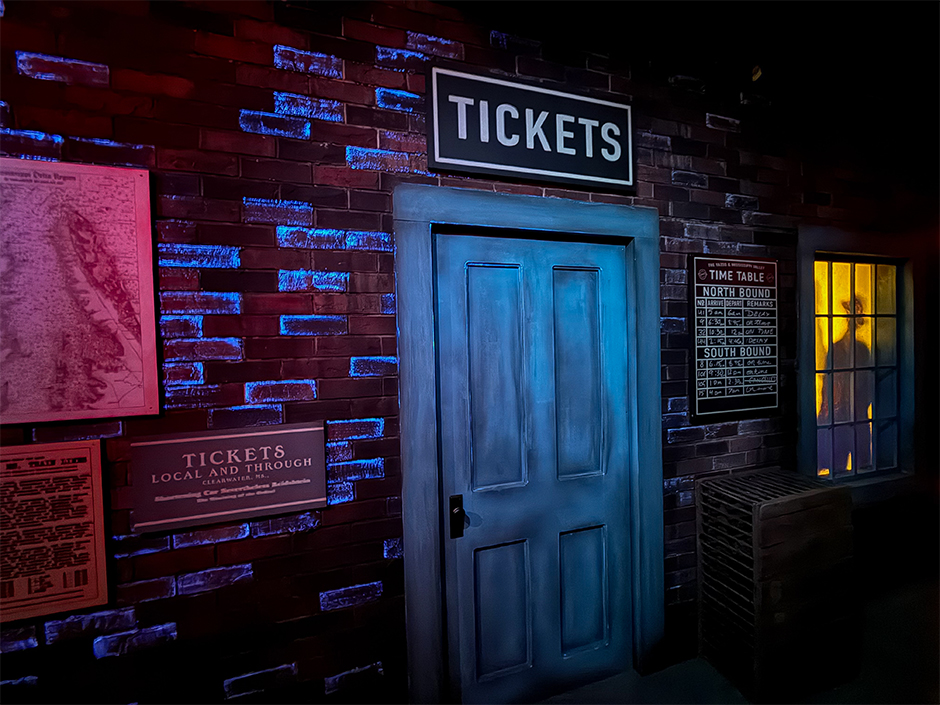 The Clarksdale train station set inside the Sinners haunted house at Halloween Horror Nights 35. A blue painted door under a TICKETS sign sits in a red brick wall lit in blue and magenta, with a hand lettered timetable board to the right and a silhouetted figure standing in a yellow lit window.