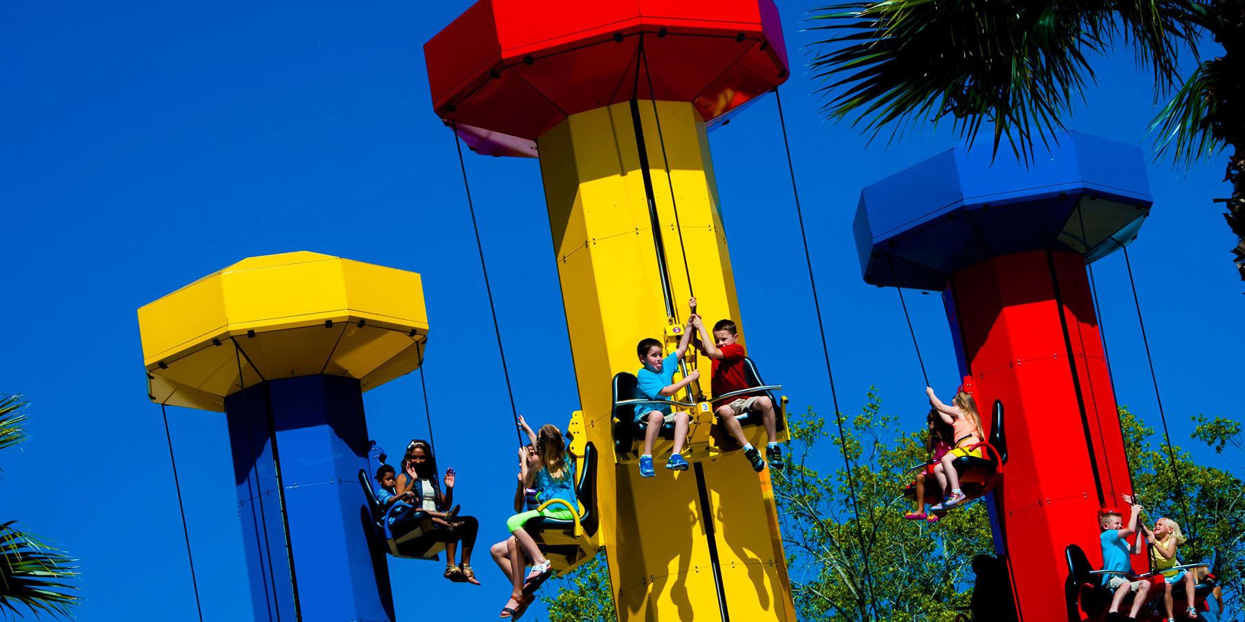 Three LEGO brick styled towers in red, yellow and blue with children seated in harnesses partway up, against a blue sky