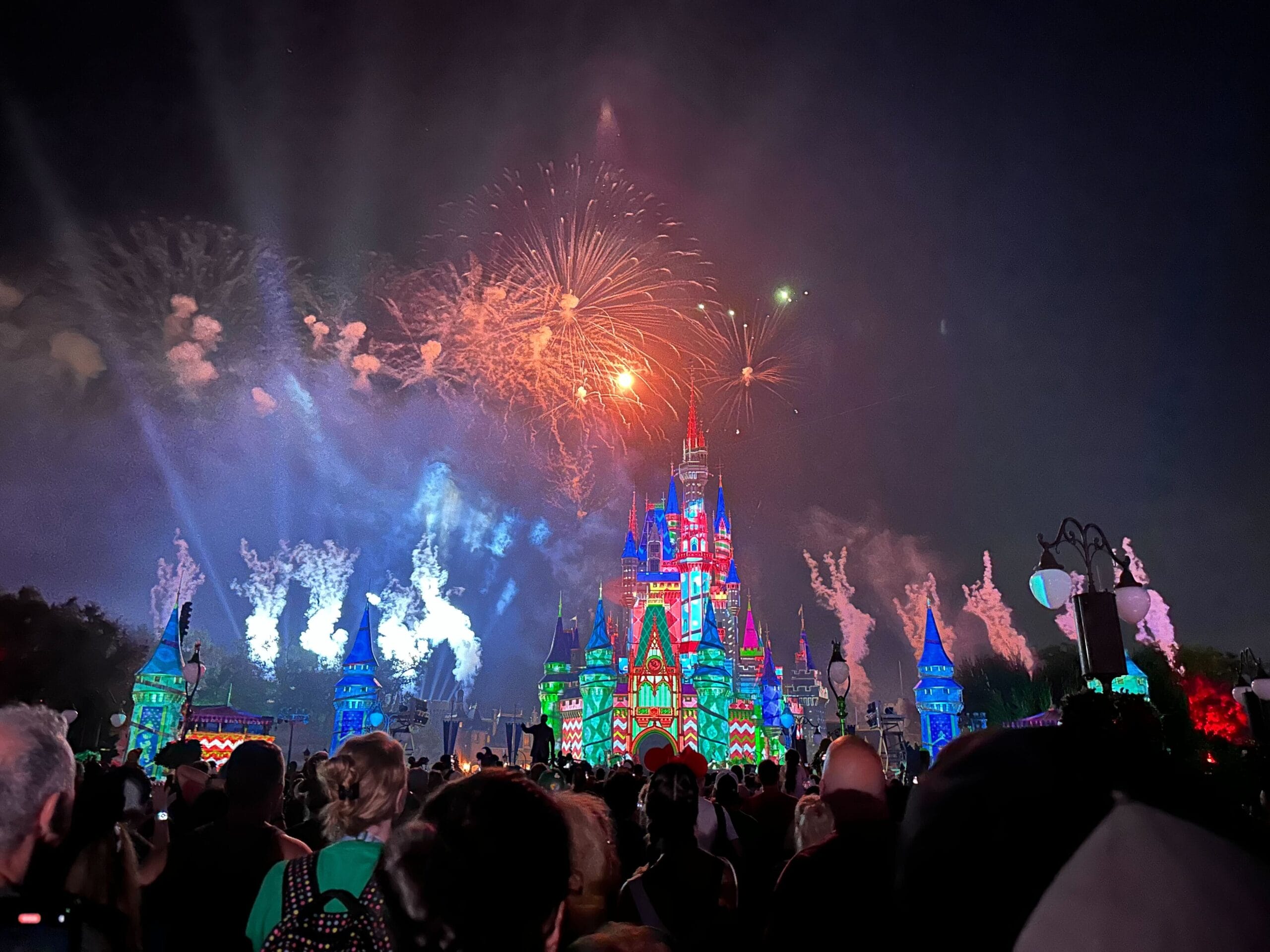 Fireworks bursting in orange and white above Cinderella Castle at night, with the castle projection mapped in red, green and blue holiday patterns and a large crowd watching in silhouette in the foreground.