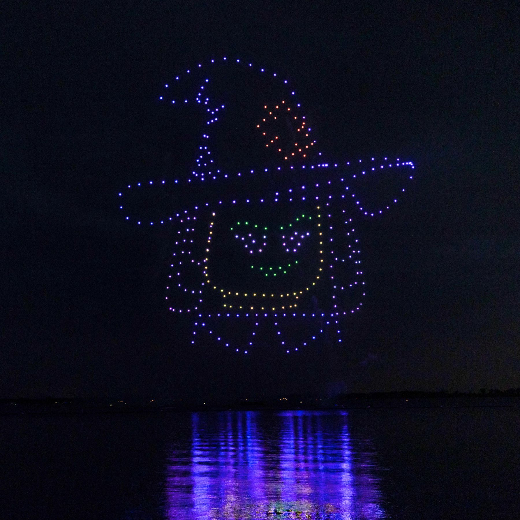 A Halloween drone show over dark water at night, hundreds of lit drones forming a smiling LEGO minifigure face wearing a witch hat, with the colored lights reflecting in the lake below.