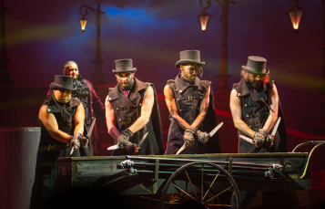Monster Stomp performers in top hats and leather vests on a Victorian street set behind a green handcart