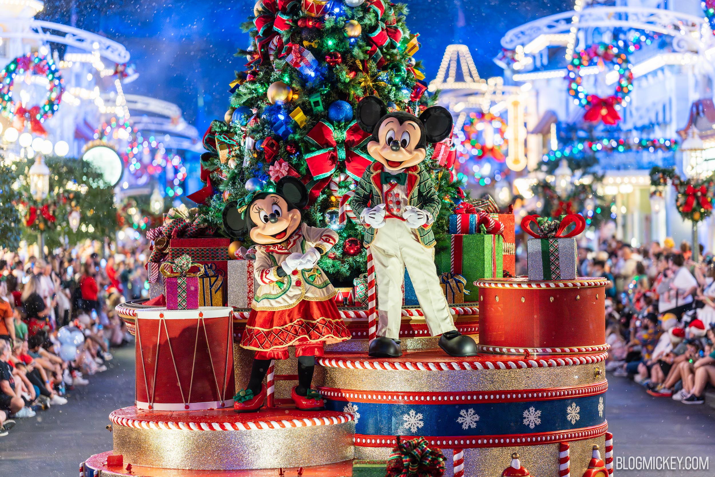 Mickey Mouse in a green plaid coat and cream trousers and Minnie Mouse in a cream jacket and red skirt posing on a decorated parade float in front of a lit and ornamented Christmas tree on Main Street U.S.A., with guests seated on both sides and artificial snow falling.