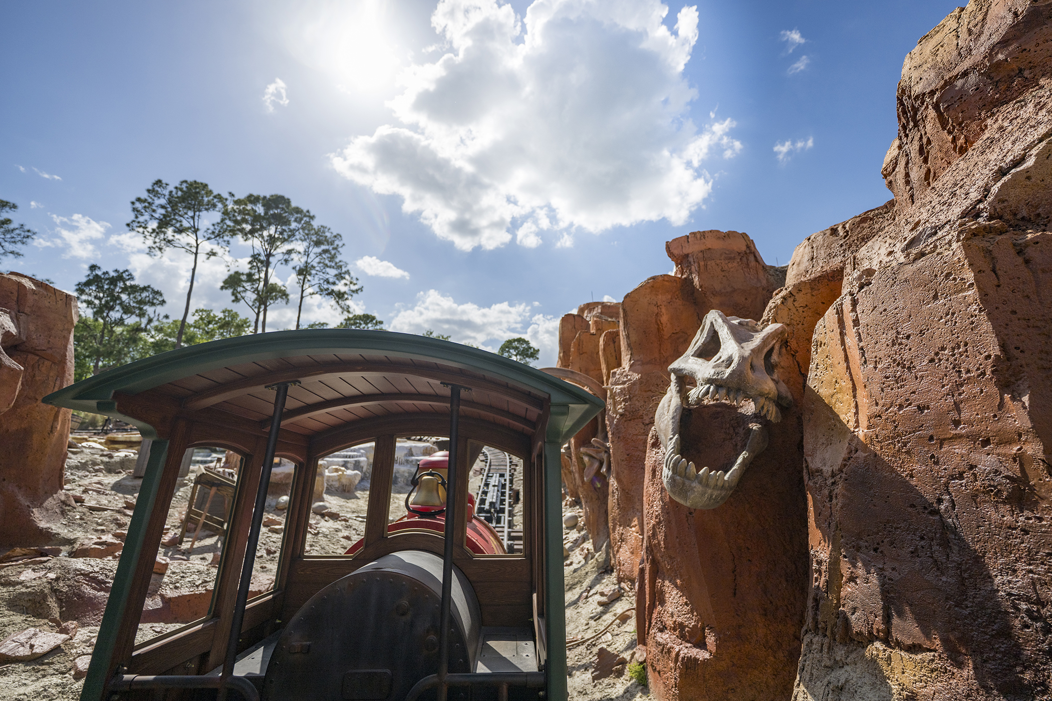 Sunlit view forward from a Big Thunder Mountain Railroad train into a red rock canyon, with a large pale fossilized skull mounted in the rock face at right, jaws open.