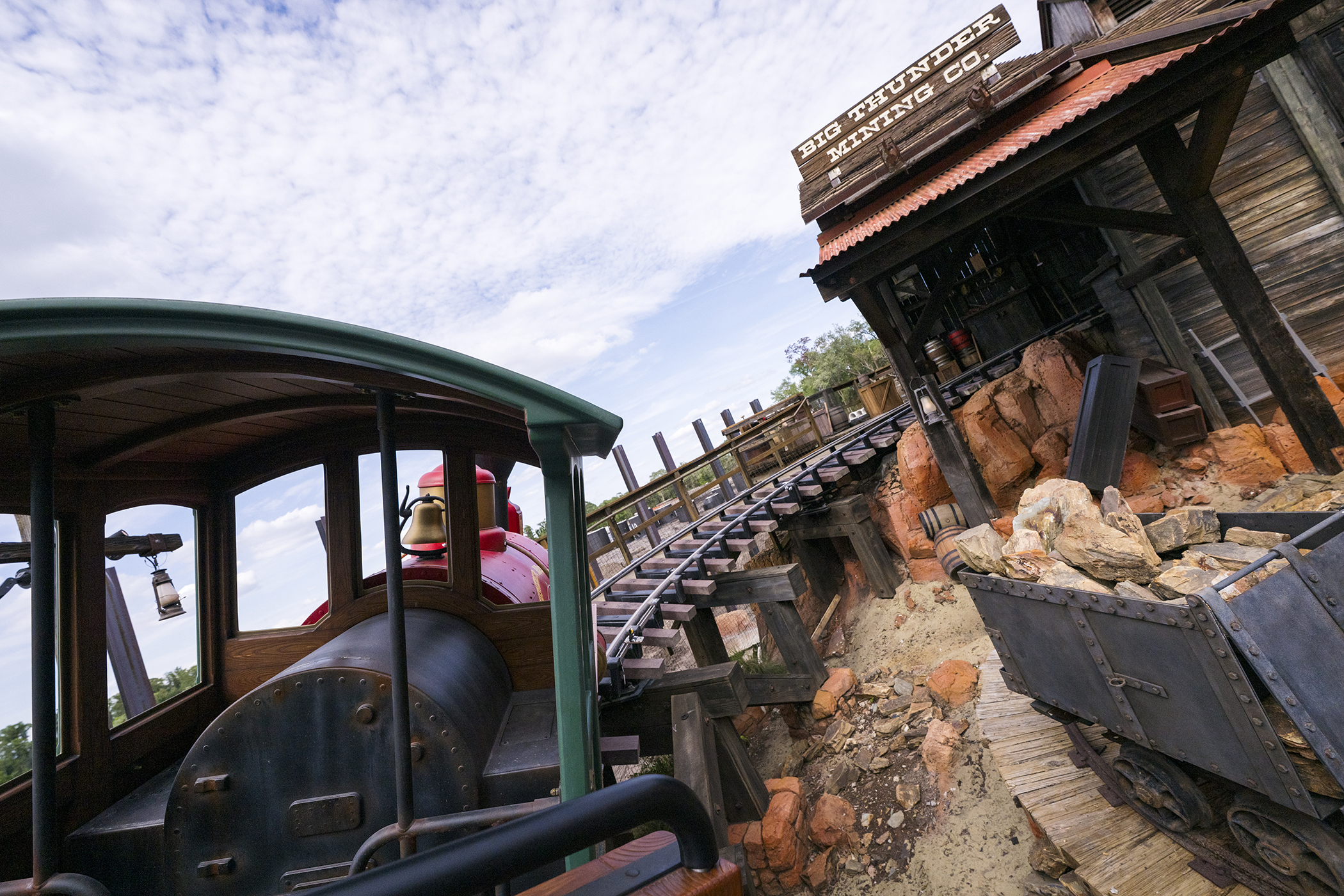 View forward from under the green canopy of a Big Thunder Mountain Railroad train, past the red boiler and brass bell, toward a weathered timber building signed BIG THUNDER MINING CO. with track and wooden crossties climbing to the right.