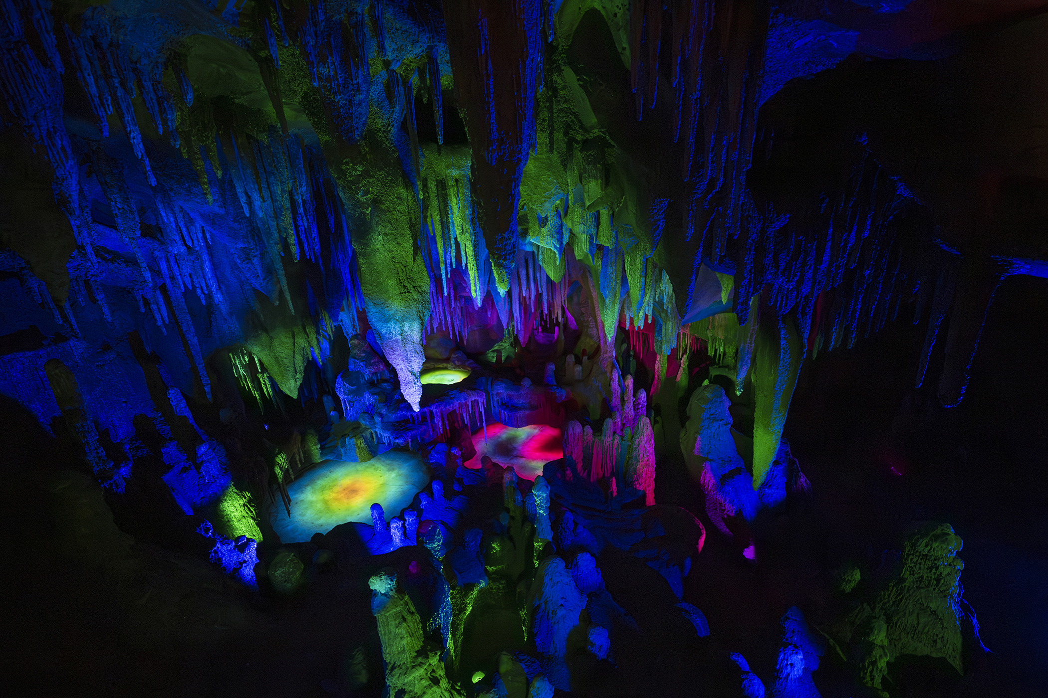 The Rainbow Caverns scene photographed in the dark, with stalactites and stalagmites lit in blue, green and magenta above glowing yellow and pink pools.