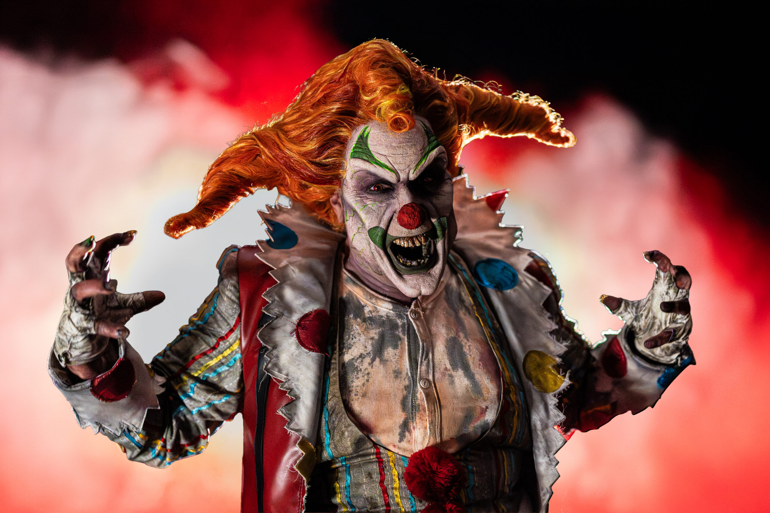Jack the Clown in a studio portrait, hands raised with long clawed fingers, mouth open in a snarl showing jagged teeth, orange jester-horn hair, cracked white face paint with green markings and a red nose, wearing a tattered polka-dot clown coat, against red-lit smoke.