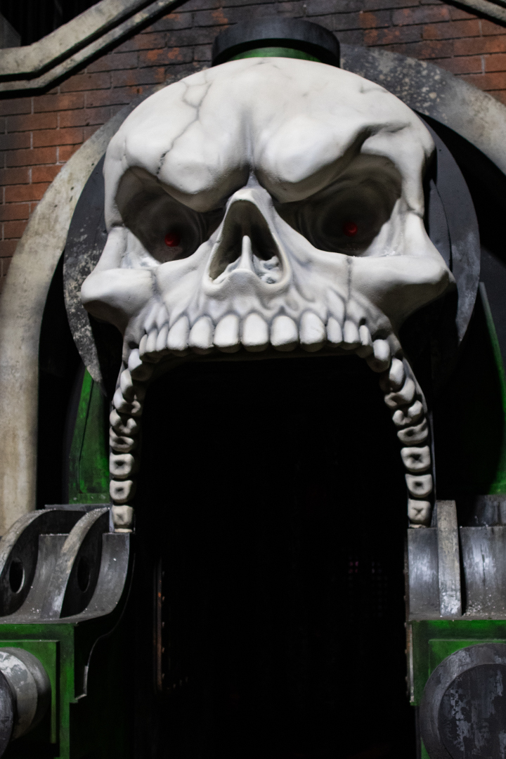 A large white skull with glowing red eyes mounted over a doorway, its open jaw and teeth forming the frame guests walk through, flanked by green painted industrial machinery and brick.