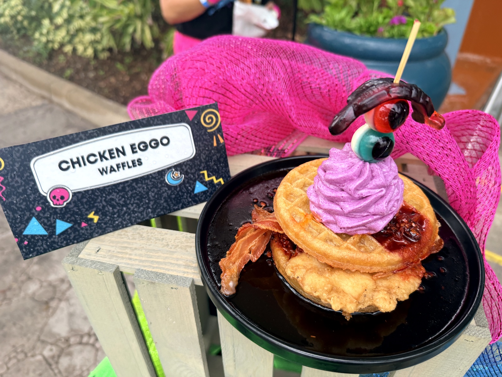 Two round waffles stacked over a fried chicken cutlet on a black plate pooled with dark syrup, a strip of bacon fanned out to the left, red jam at the edge, and a swirl of bright purple whipped cream on top skewered with a gummy eyeball and a dark gummy worm. A black card at left reads CHICKEN EGGO WAFFLES.