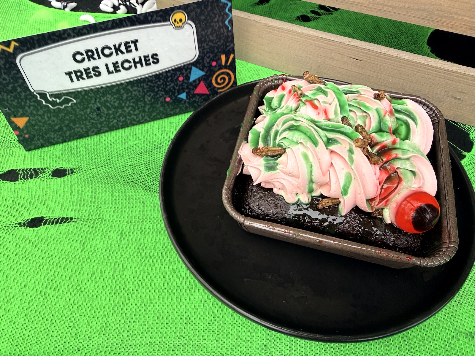 A rectangular foil tray of dark soaked sponge cake topped with thick swirls of pink and green piped frosting drizzled with red syrup and studded with several whole roasted insects, plus a red and black gummy eyeball at one corner, sitting on a black plate on bright green mesh. A black card at left reads CRICKET TRES LECHES.