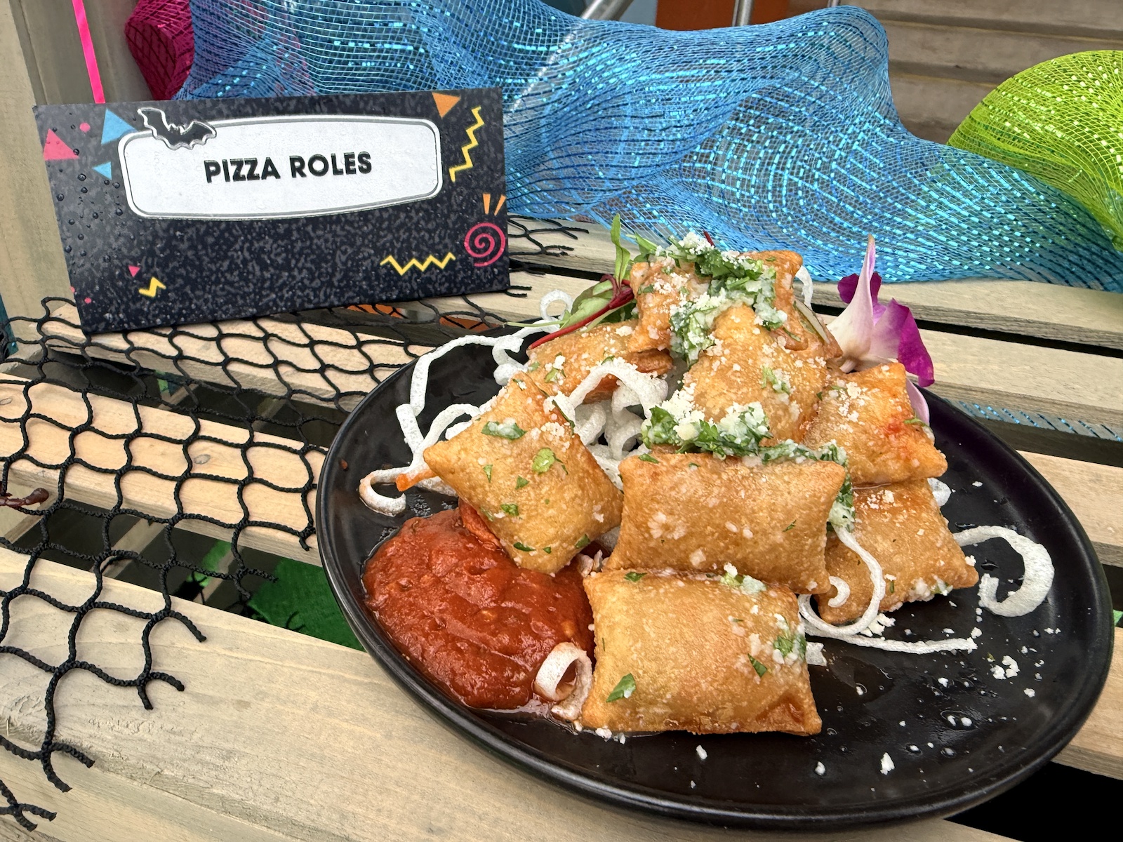 A stack of golden fried pizza rolls on a black plate over a nest of crisp white fried curls, dusted with crumbled white cheese and chopped herbs, with a pool of red marinara at the left and a purple orchid garnish. A black card at upper left with a bat icon and neon squiggles reads PIZZA ROLES.