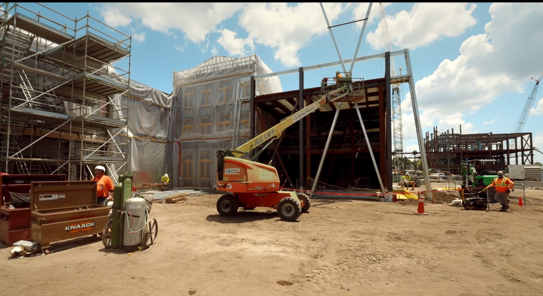 Monstropolis construction site with scaffolding at left, a plastic-wrapped multi-story facade with arched windows, raw steel framing at right, an orange boom lift in the foreground and lattice-boom cranes behind