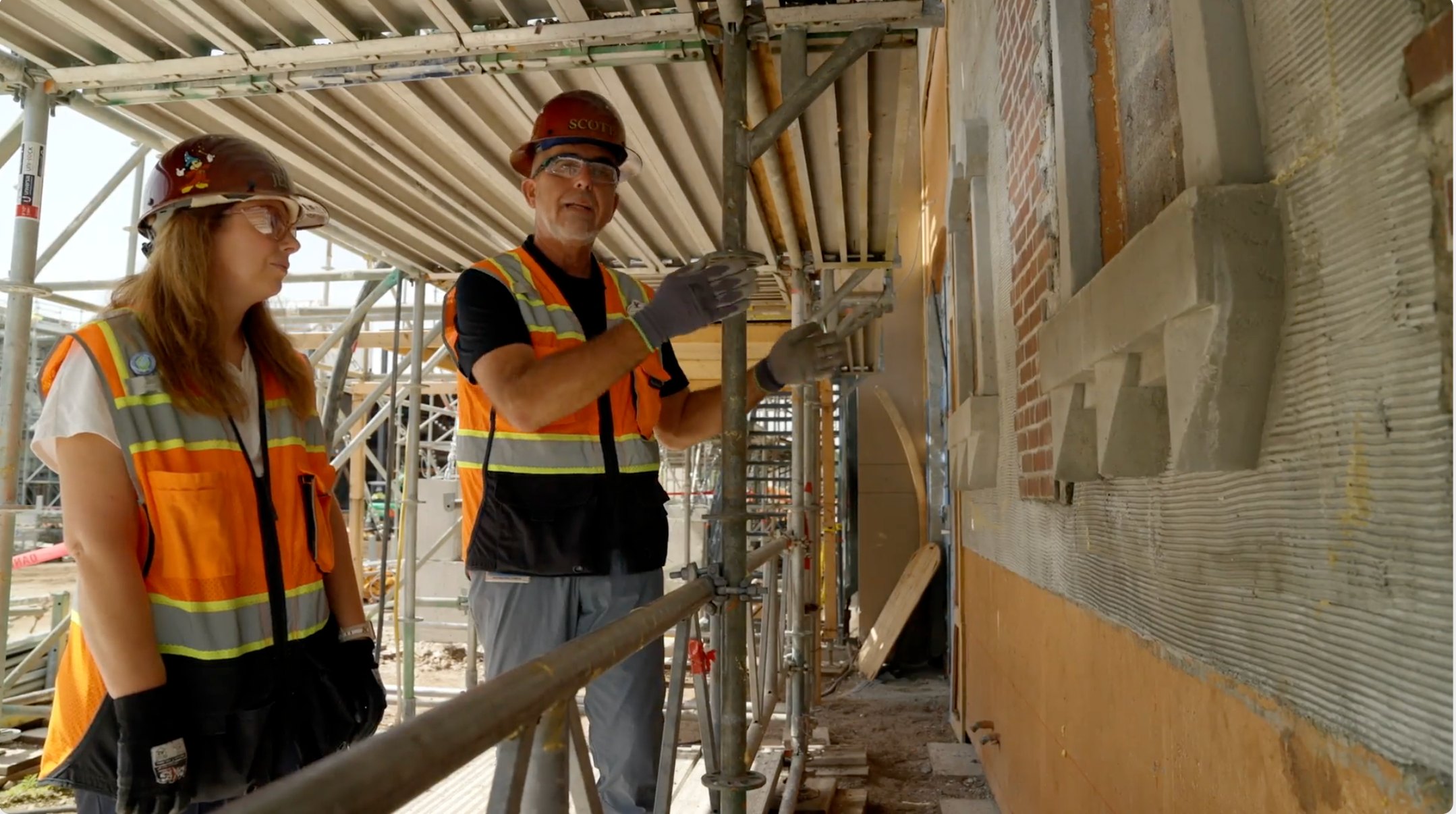 Two Imagineers in hard hats and orange safety vests standing on scaffolding beside a partly finished wall showing exposed brick, ribbed grey scratch coat and a cast stepped moulding