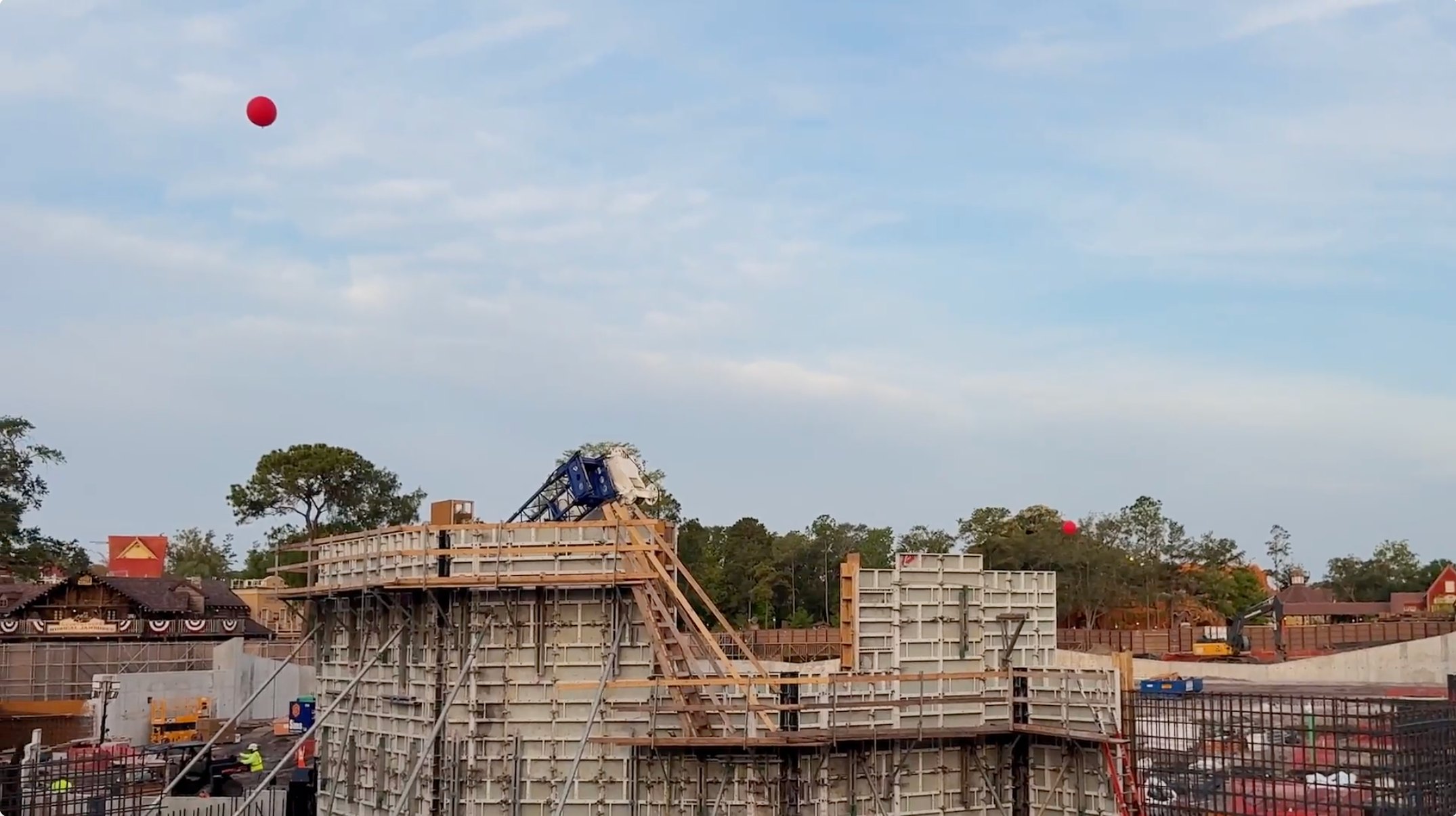 Two red test balloons at different heights above the Piston Peak construction site, with concrete formwork, the blue lattice boom of a crane and the Country Bear Musical Jamboree building visible