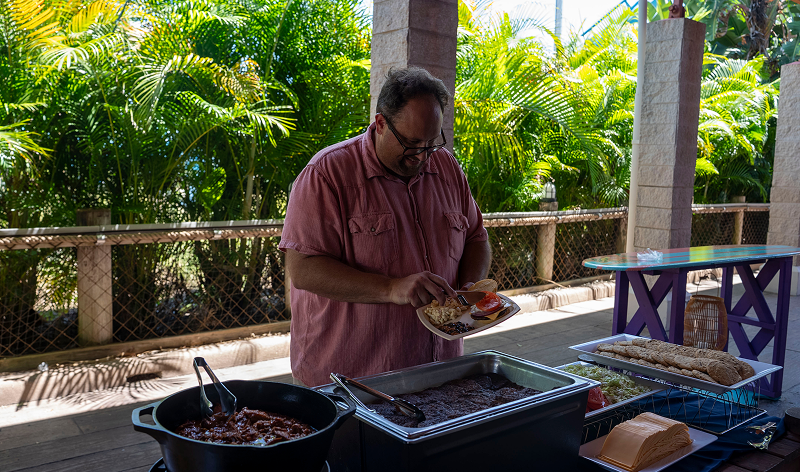 A guest filling a plate from a covered outdoor buffet line of chafing pans and a tray of rolls