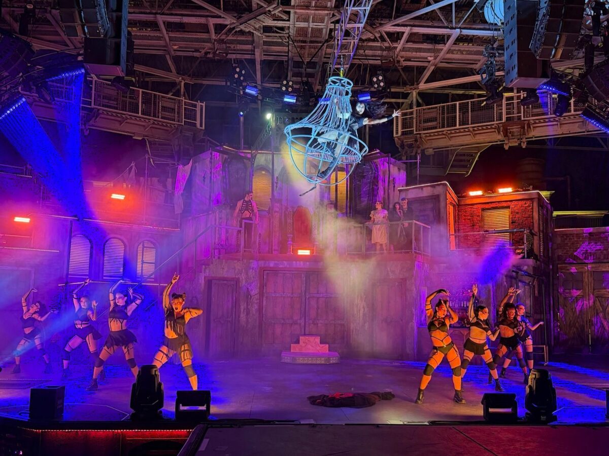 The Blood Noir stage lit in purple and orange. A performer in dark costume is suspended high in the center inside a large open framework hanging from the rigging, shaped like an inverted bell of pale curved bars. Two groups of about five dancers each are posed in harness costumes on the stage floor to the left and right with arms raised, a shape resembling a body lies flat center stage, and the tenement set with shuttered windows and a brick building rises behind them.