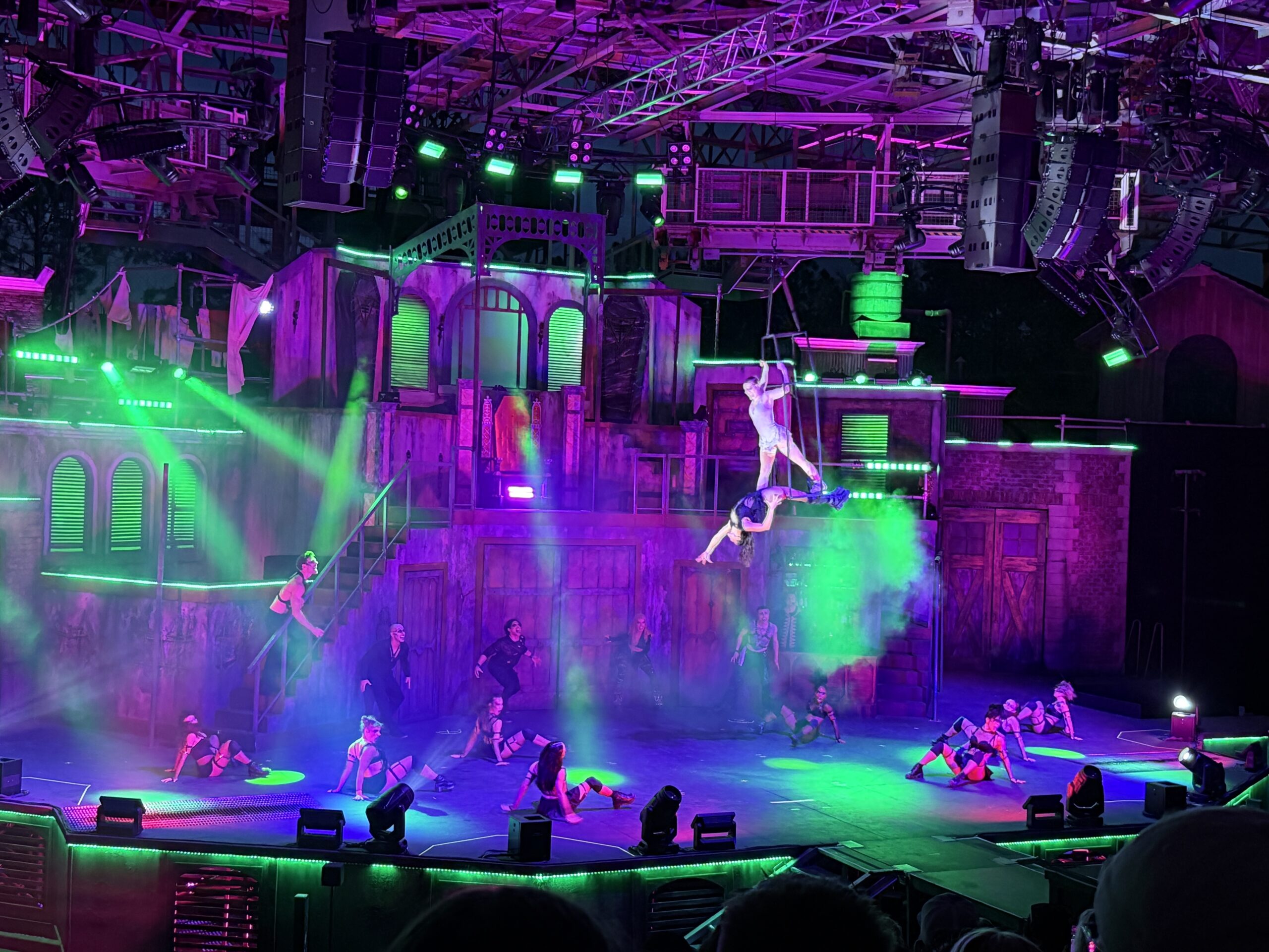 Wide view of the Blood Noir stage lit in purple and green. A multi level urban tenement set fills the background with shuttered arched windows, staircases, laundry strung on lines and a green lit water tower. One performer hangs from a vertical rig high at the right with a second performer inverted just below them in an aerial duo. Around ten dancers are low on the stage floor in crouched poses and several more stand on the raised level of the set behind them, with lighting truss and speaker stacks overhead and audience heads in silhouette along the bottom.