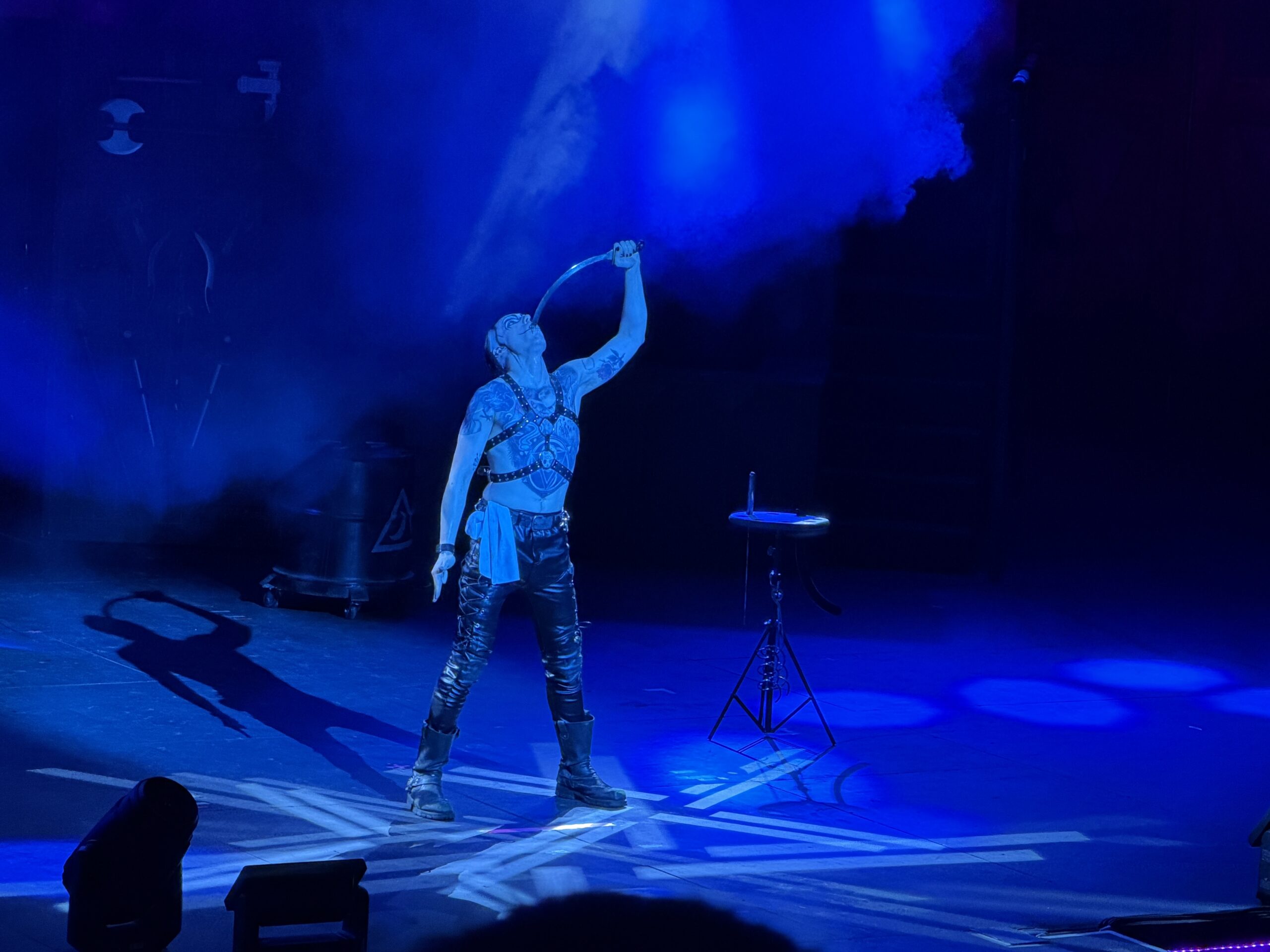 A tattooed performer stands center stage bathed in deep blue light and haze, wearing a studded black leather harness, black leather trousers and heavy boots, with a pale sash at the hip. His head is tilted fully back and he is lowering a long curved blade down into his throat, gripping the handle above him with one raised arm. A small stand holding chains and more blades sits beside him, and his shadow is thrown across the stage floor.
