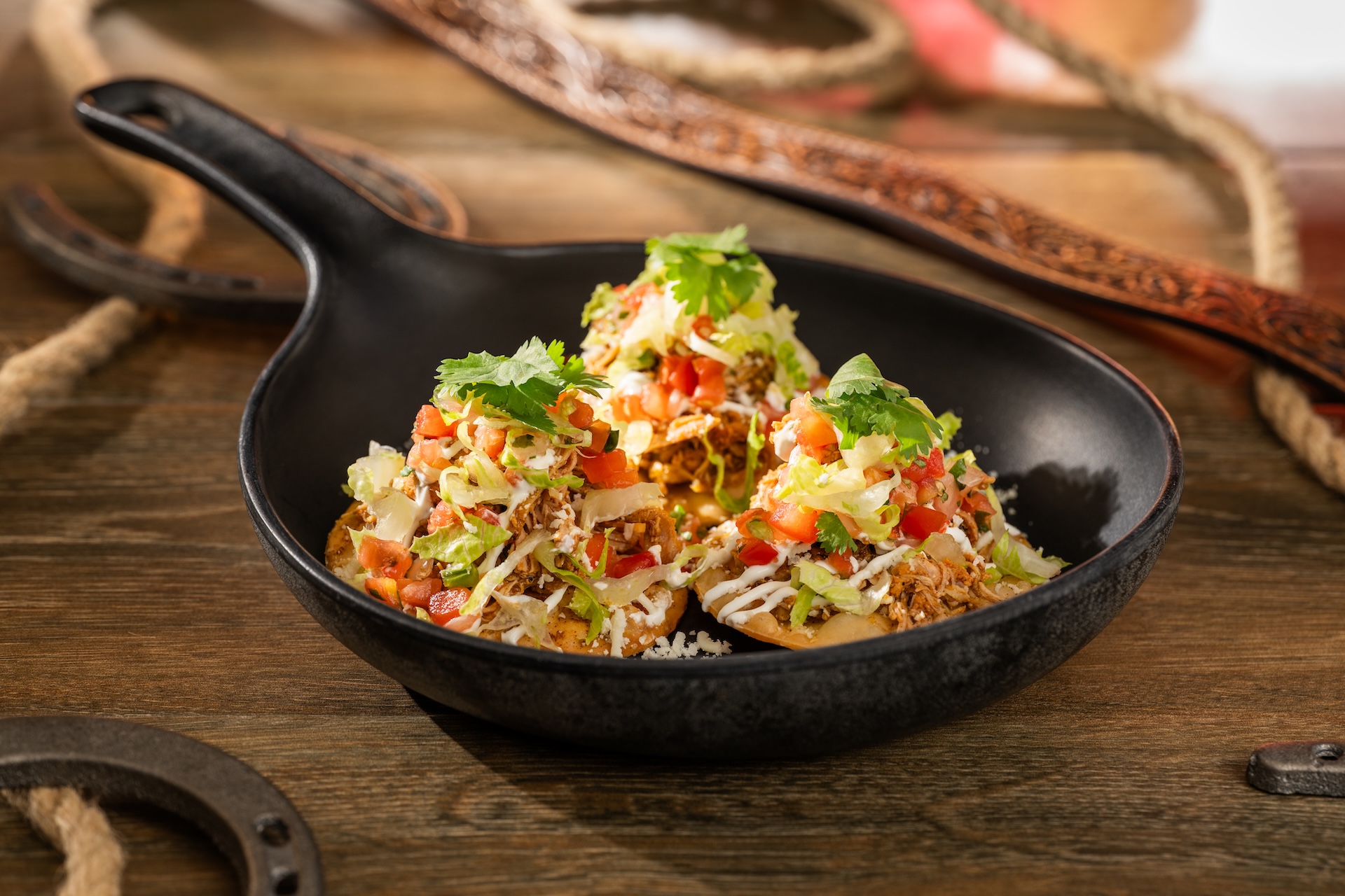 Three small tostadas topped with shredded chipotle chicken, lettuce, diced tomato, crema and cilantro, served in a black cast iron pan