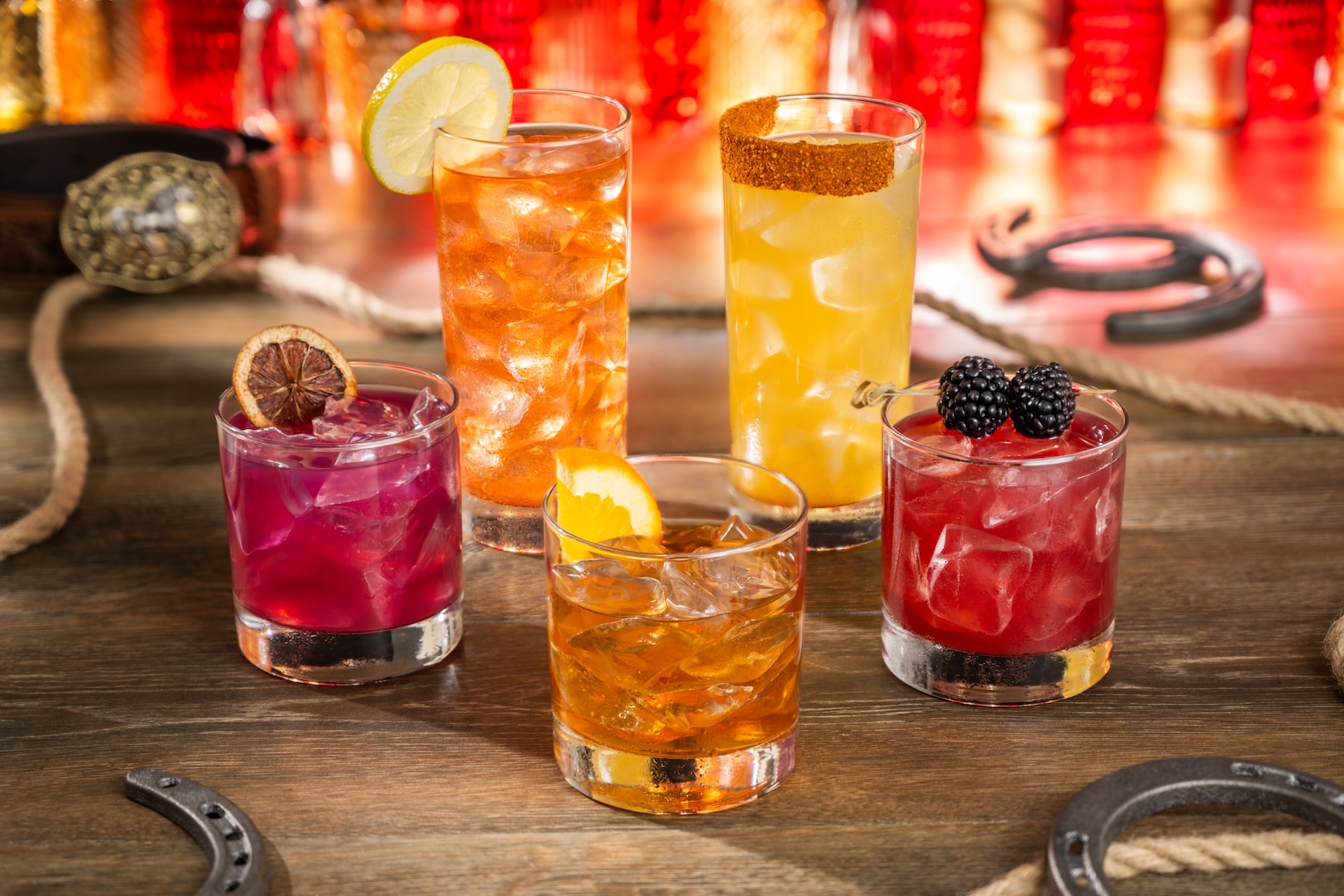 Five cocktails on a saloon table: a magenta margarita with a dried lime wheel, an orange highball with a lemon slice, a chili rimmed yellow highball, an old fashioned with an orange slice, and a red drink garnished with blackberries