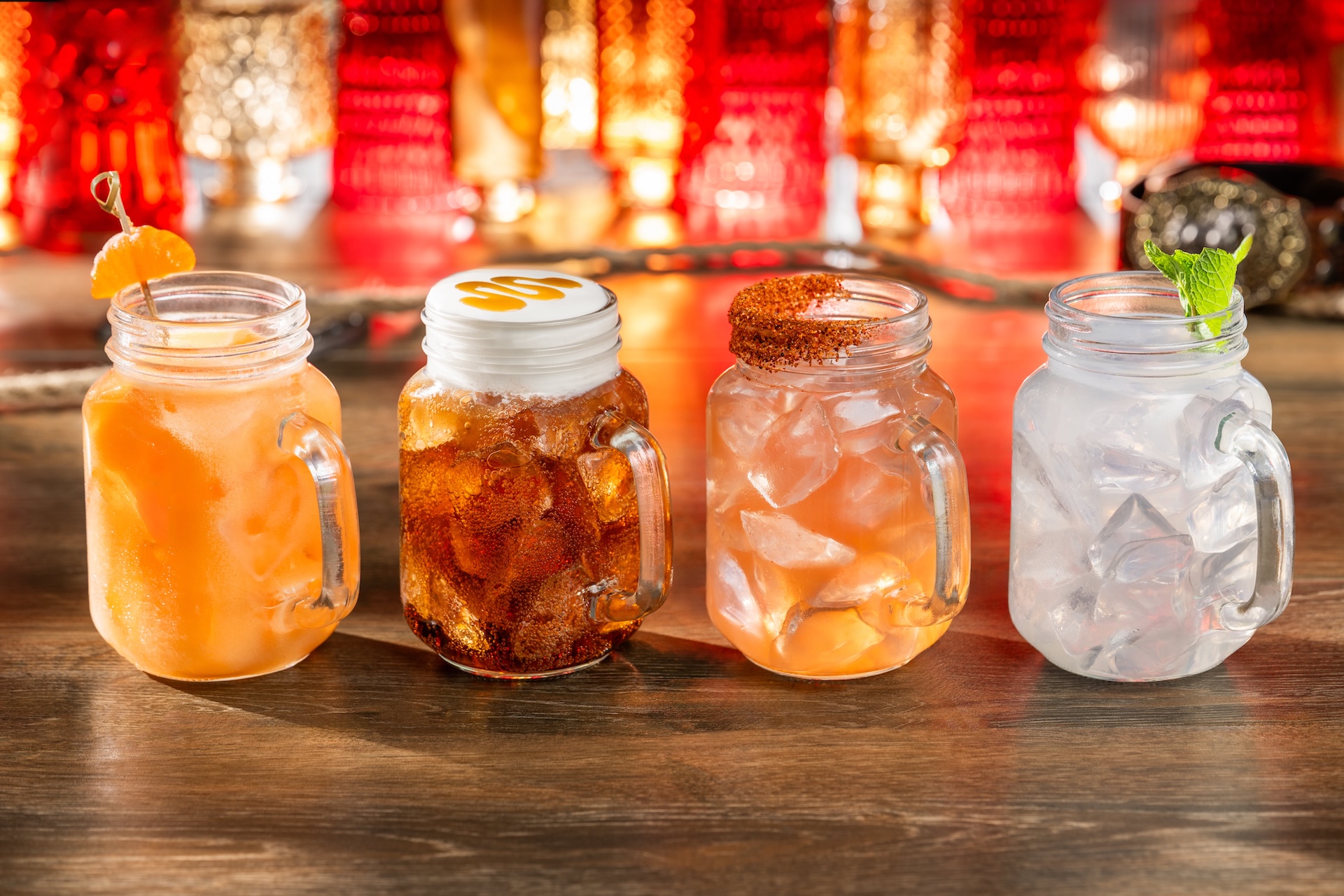 Four mason jar mugs of non alcoholic drinks on a wooden table: an orange cream soda, a root beer capped with sweet cream, a chili rimmed lemonade and a lime splash garnished with mint