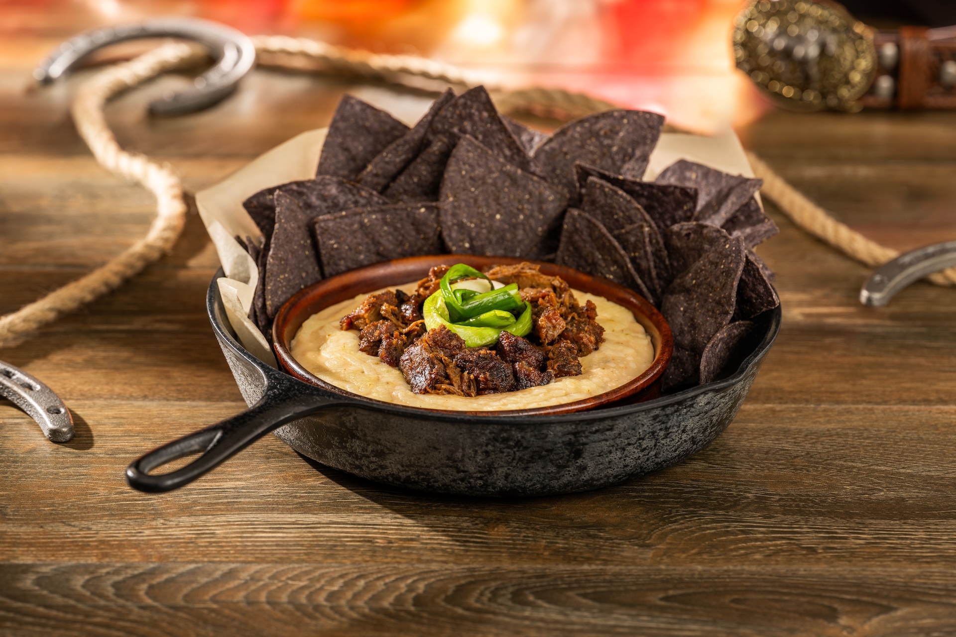 A cast iron skillet holding a bowl of queso topped with chopped barbecue burnt ends and scallions, ringed with blue corn tortilla chips