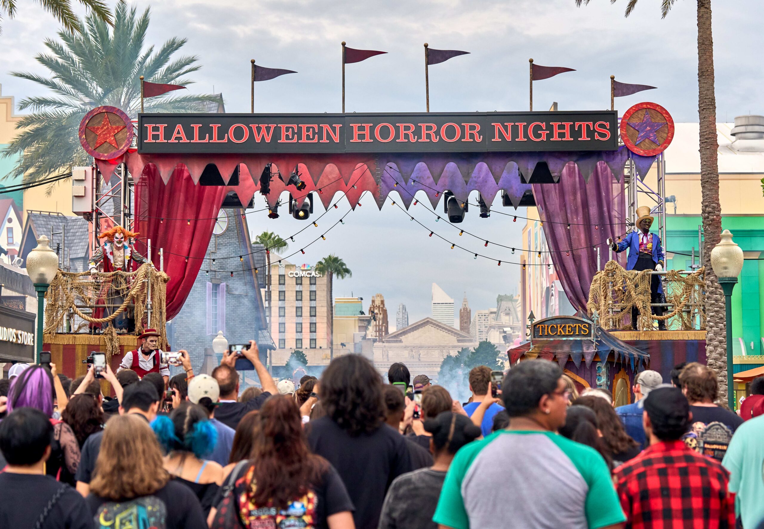 A crowd photographing the Halloween Horror Nights entrance marquee in daylight under a bright overcast sky, with a red and purple carnival valance, pennant flags, and costumed performers on raised platforms at either side, a ticket booth below the right platform