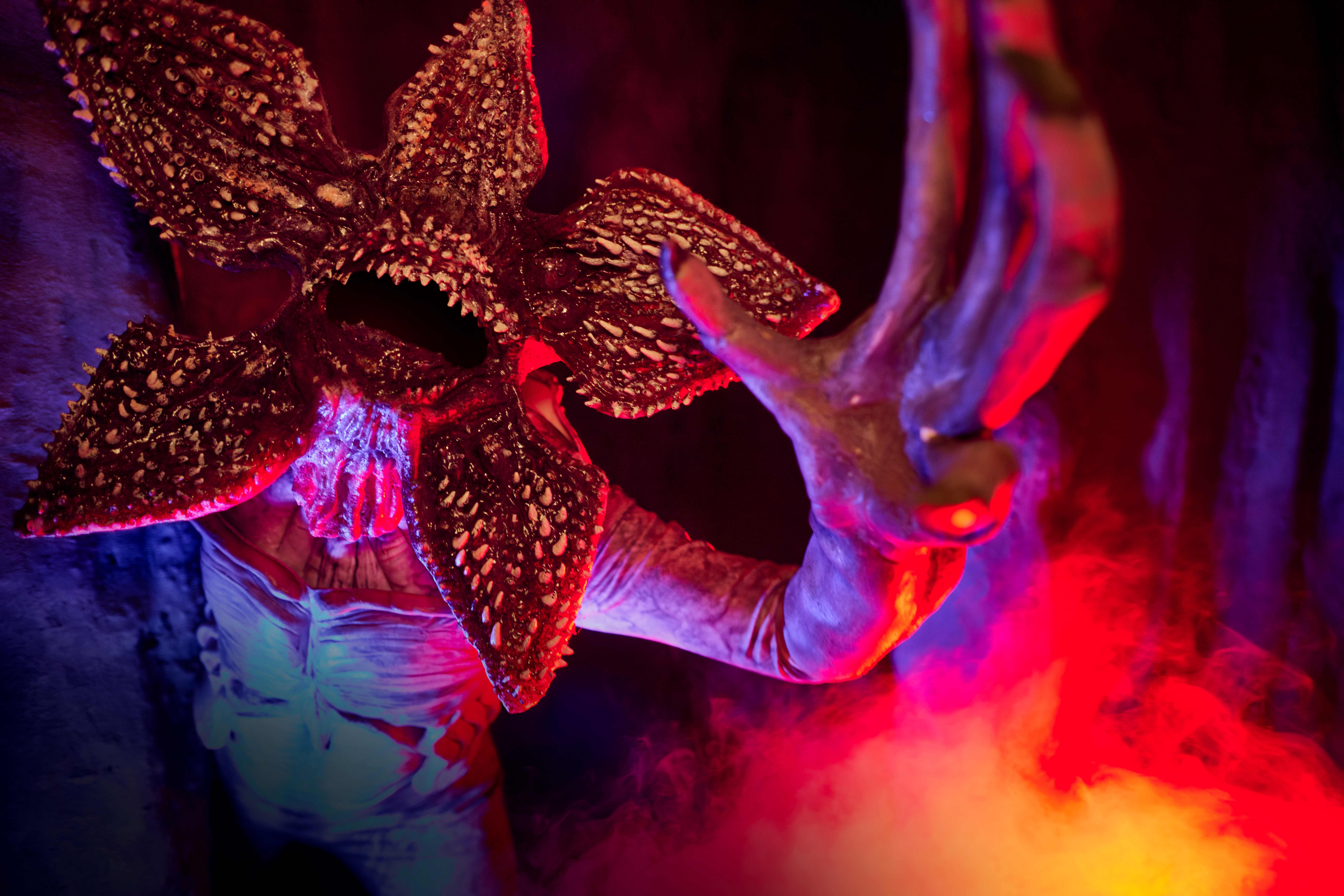 A Demogorgon in close up with its petal like head split fully open to show rings of teeth, one long clawed arm reaching across the frame, lit in red and blue with fog rolling in at the lower right