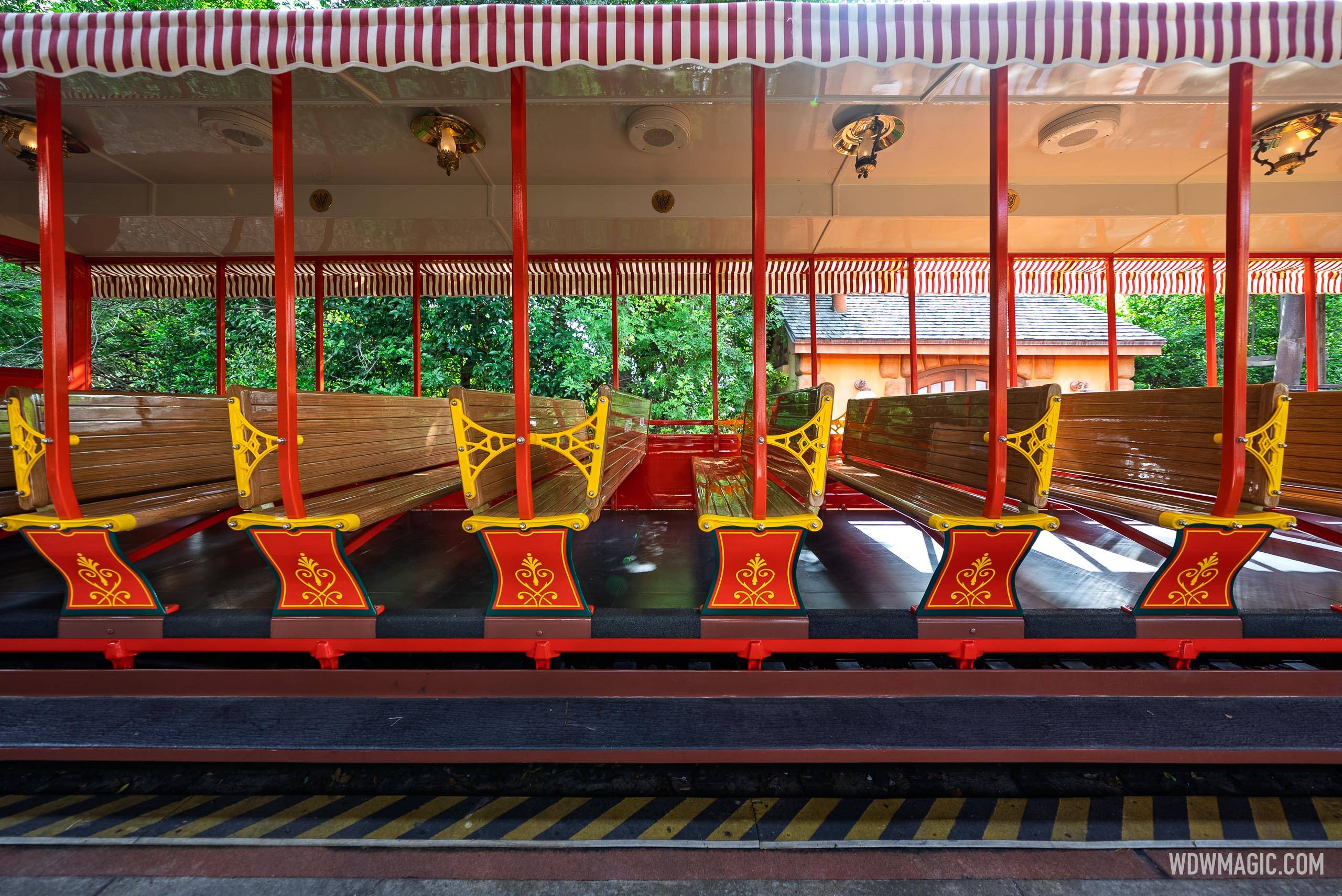 A straight on side view from the platform into an open sided railroad coach with varnished wooden bench seats on yellow cast iron frames, red support posts and a red and white striped awning
