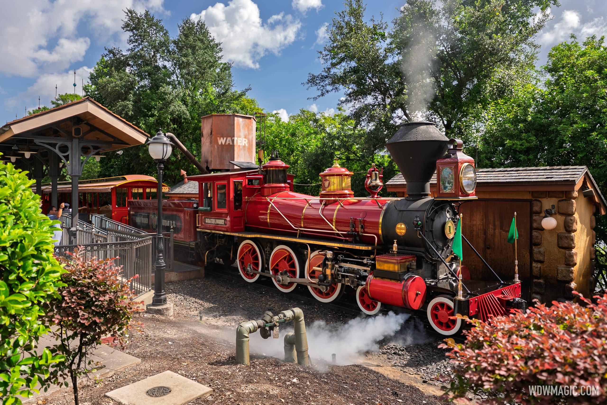The red Walter E. Disney locomotive, engine number 1, stopped just beyond a station platform with its coaches still alongside it, venting steam near the track, and a rusted water tank behind