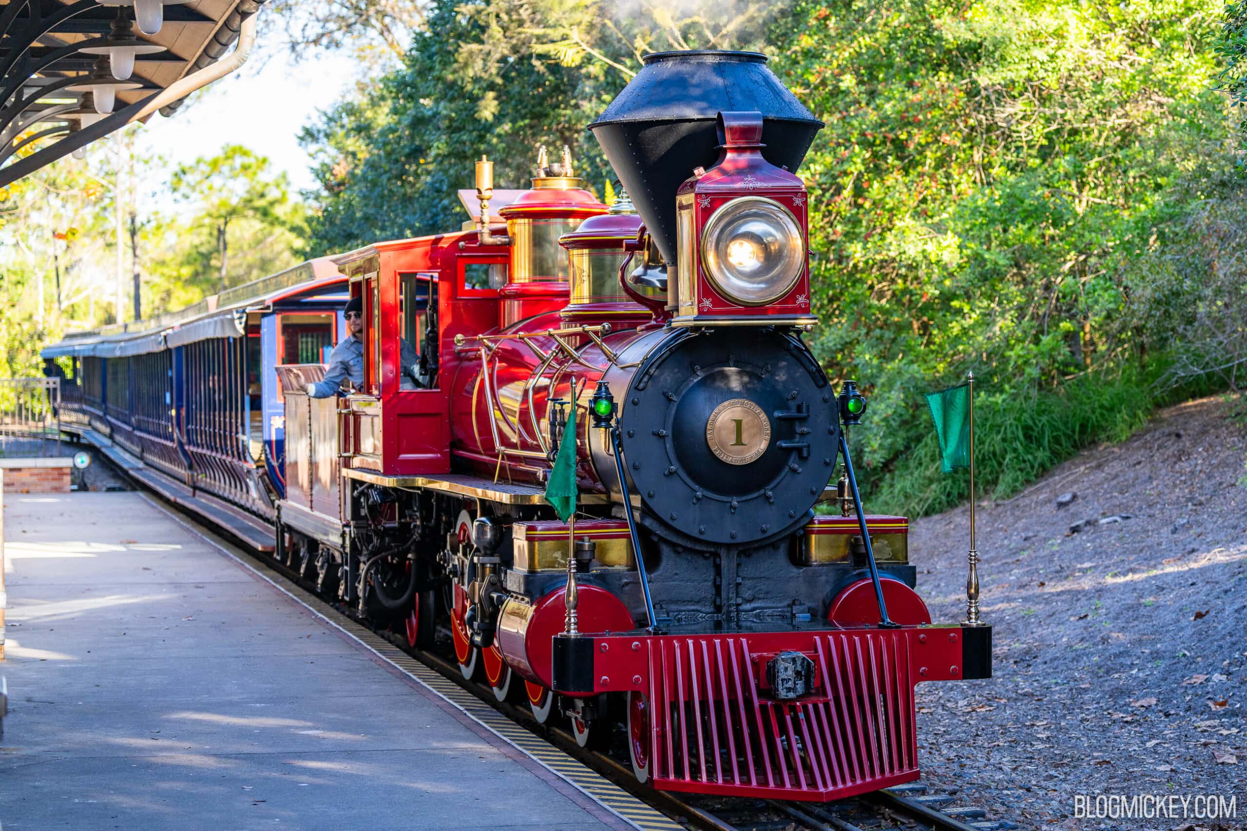 A red Walt Disney World Railroad steam locomotive carrying engine number 1 on its smokebox door, headlight lit and an engineer leaning from the cab, pulling a line of dark blue open sided coaches alongside a station platform