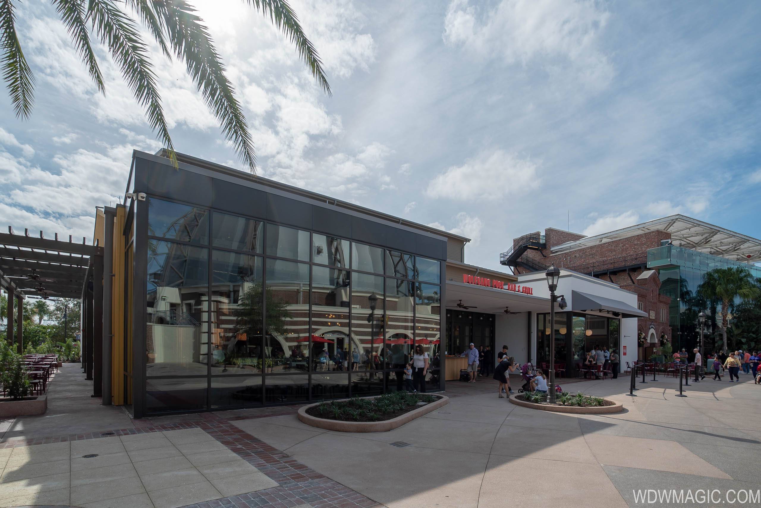 Exterior of Wolfgang Puck Bar & Grill at Disney Springs on a bright day, a glass walled corner dining room with dark metal framing at left, the red WOLFGANG PUCK BAR & GRILL sign mounted above the entrance canopy at right, palm fronds overhanging the top of the frame and guests walking the pavement outside.