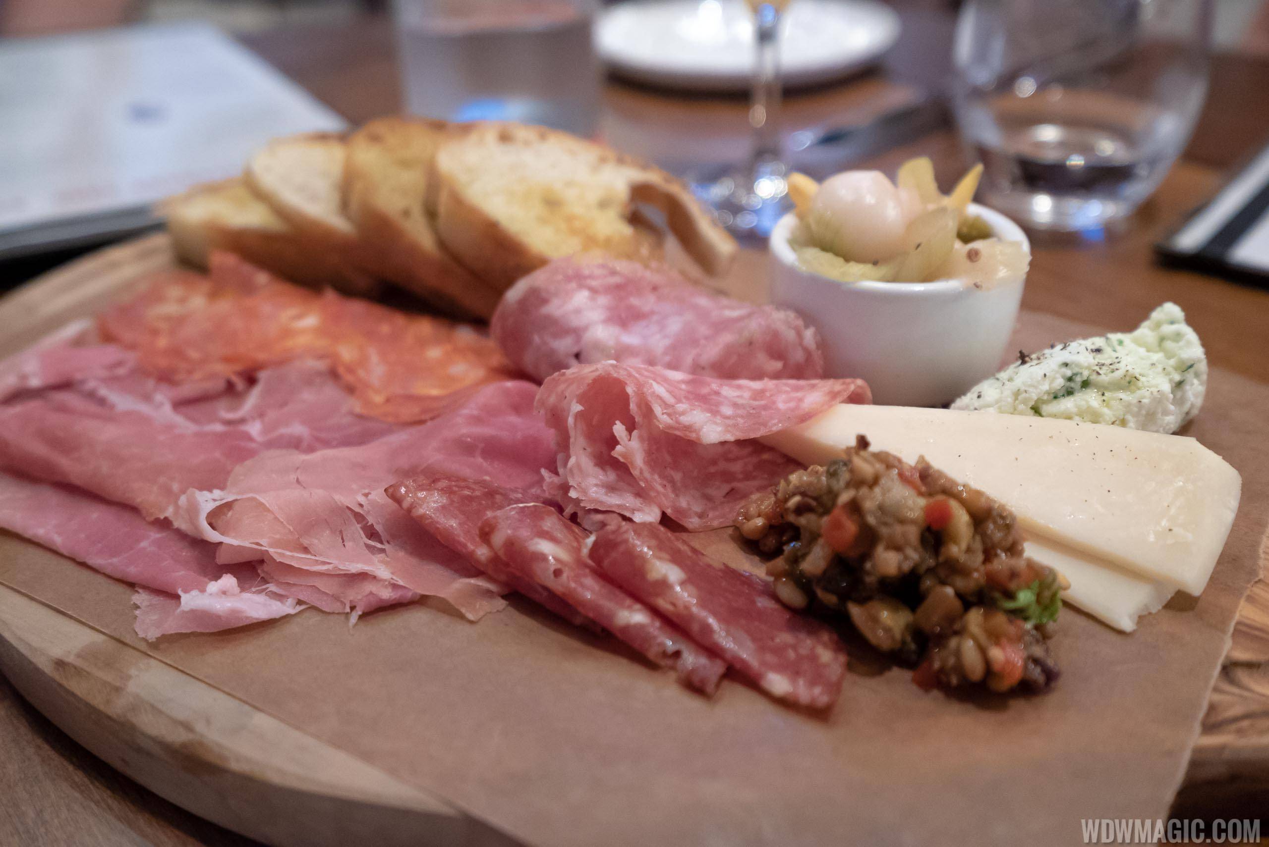 A round wooden board on a restaurant table holding folded slices of prosciutto, salami and coppa, three slices of toasted bread at the back, a wedge of pale hard cheese and a quenelle of herbed white cheese dusted with pepper at right, a spoonful of chunky dark olive relish in front of them and a small white ramekin of pickled vegetables with a cocktail onion at the top right, with water glasses out of focus behind.