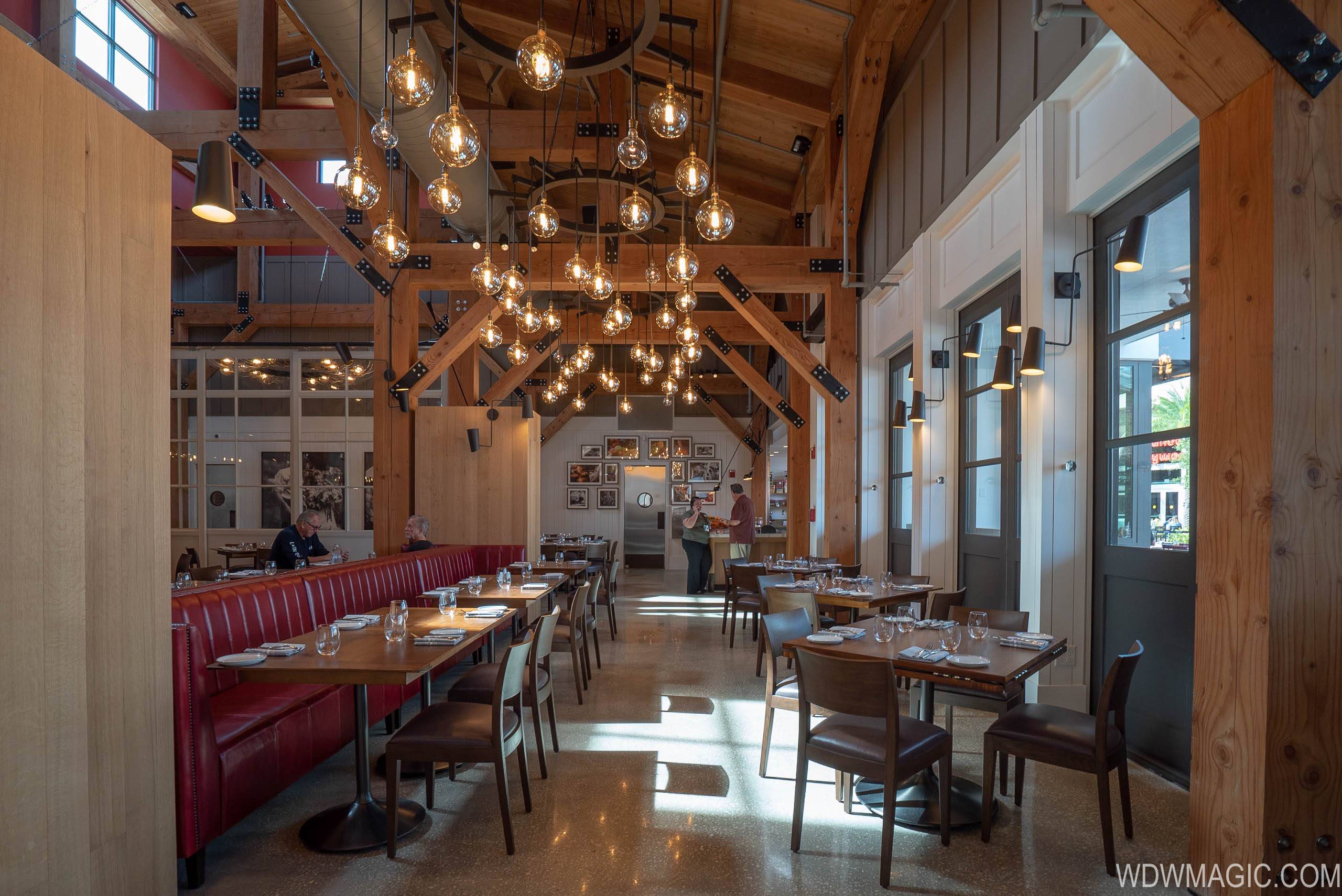 Interior dining room at Wolfgang Puck Bar & Grill, with exposed pale timber trusses and steel plate joinery overhead, dozens of clear glass Edison bulbs hanging at varied heights from two circular chandelier rings, a long red leather banquette running down the left side behind wooden tables set with wine glasses, brown leather chairs at right and a polished terrazzo floor.