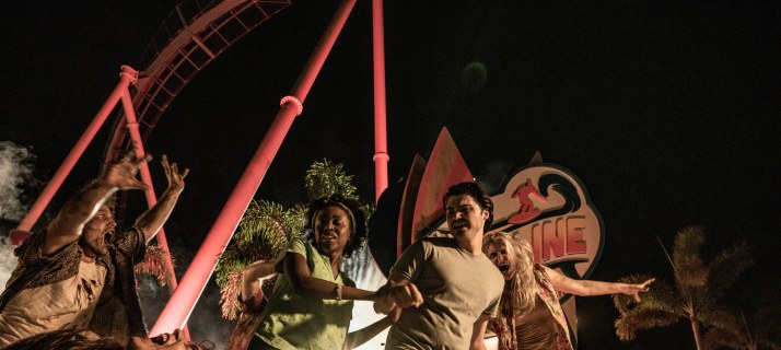 Night scene with guests reacting to costumed scare actors in ragged clothing, the red lit track of the Pipeline coaster and the illuminated Pipeline surfer sign behind them
