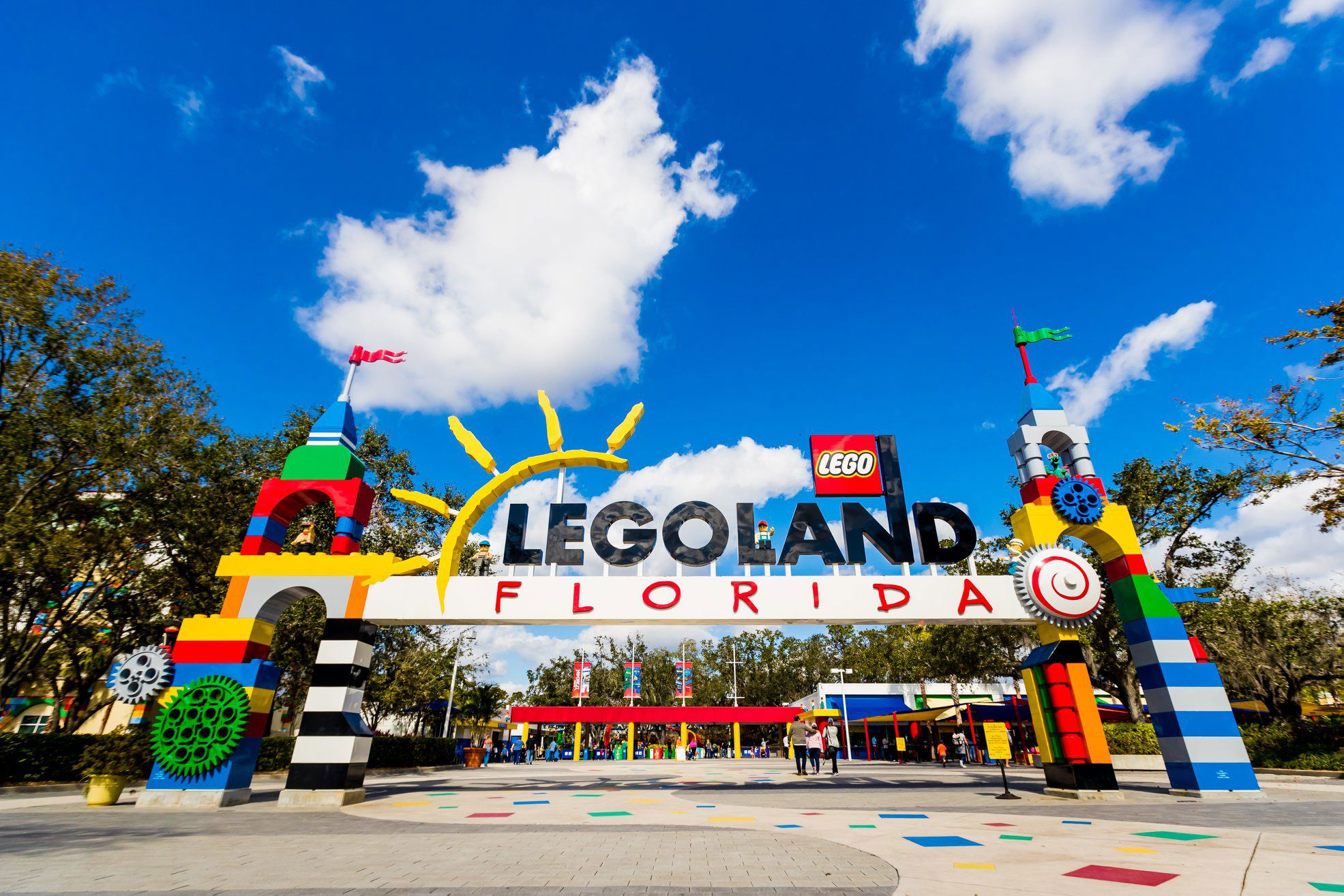 A wide panoramic view of the Legoland Florida main entrance, its arch built from oversized colored bricks spelling out Legoland above a white band reading Florida, flanked by multicolored brick towers with flags. A few guests stand at the turnstiles beneath the arch and the plaza pavement is inlaid with colored brick shapes, under a bright blue sky with wispy clouds