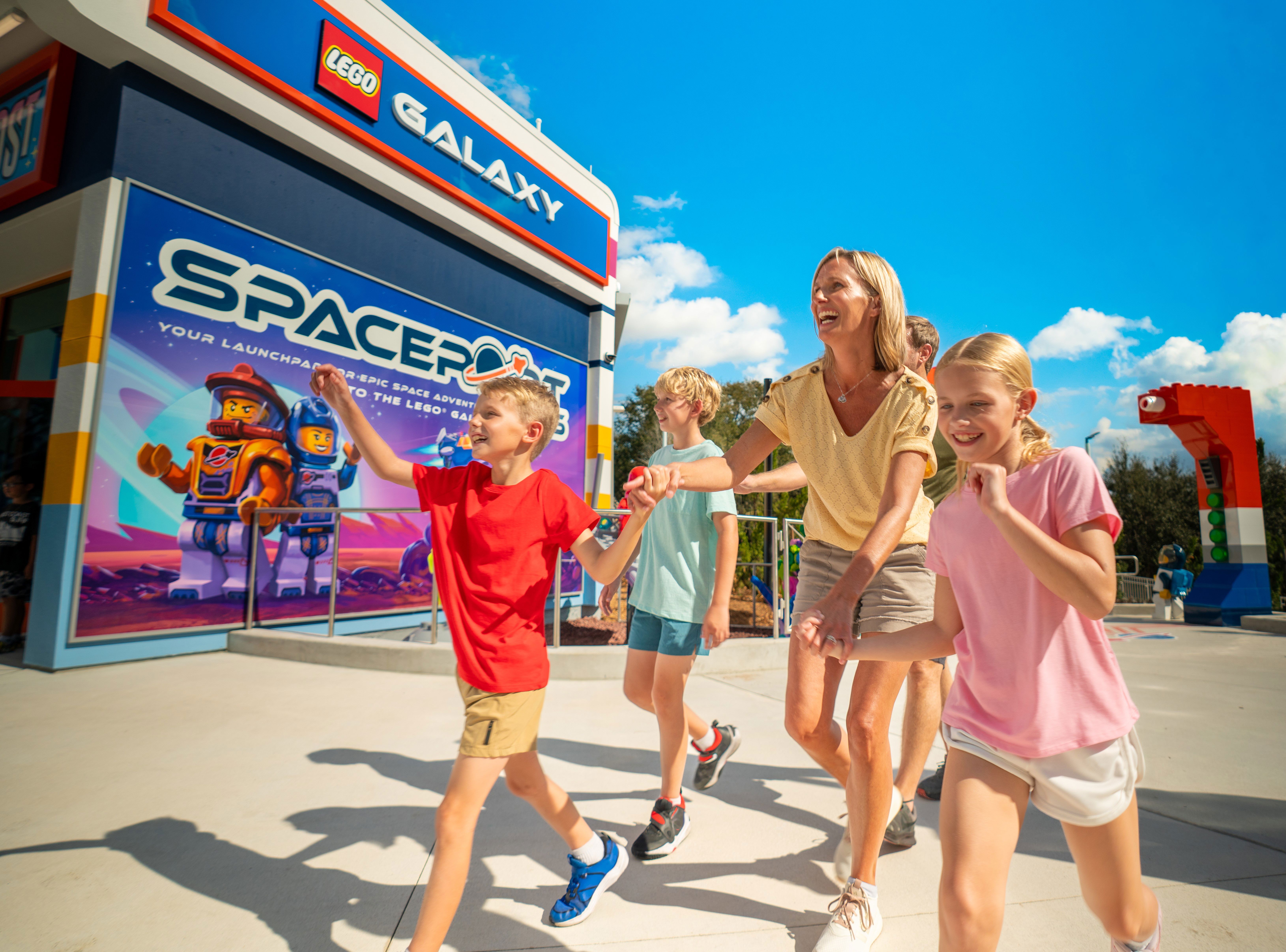 A woman, a man and three children walking away from the LEGO Galaxy building at Legoland Florida, its facade carrying a navy blue GALAXY sign in white capitals beside a red LEGO logo tile, above a large Spaceport mural showing two LEGO astronaut minifigures, one in an orange suit and one in blue, standing on purple rocky ground, under a deep blue sky