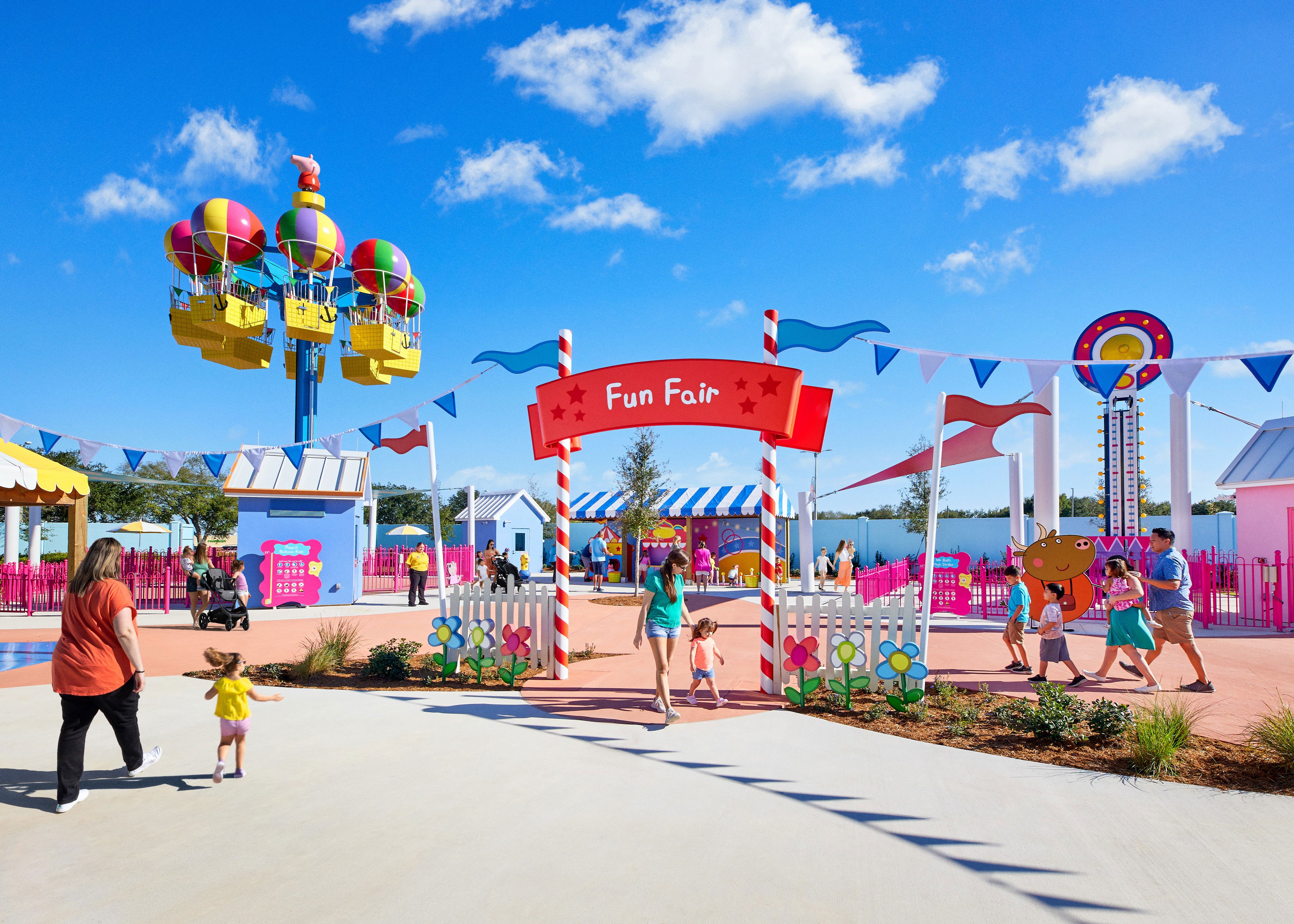 The Fun Fair area at Peppa Pig Theme Park Florida with a red Fun Fair archway on red and white striped poles above a white picket fence set with cutout flowers, a balloon themed spinner ride at left carrying pink, purple, green, yellow and red balloon canopies over yellow baskets, a high striker tower topped by a bullseye target at right, hot pink metal fencing, blue and white bunting and families walking the path under a blue sky