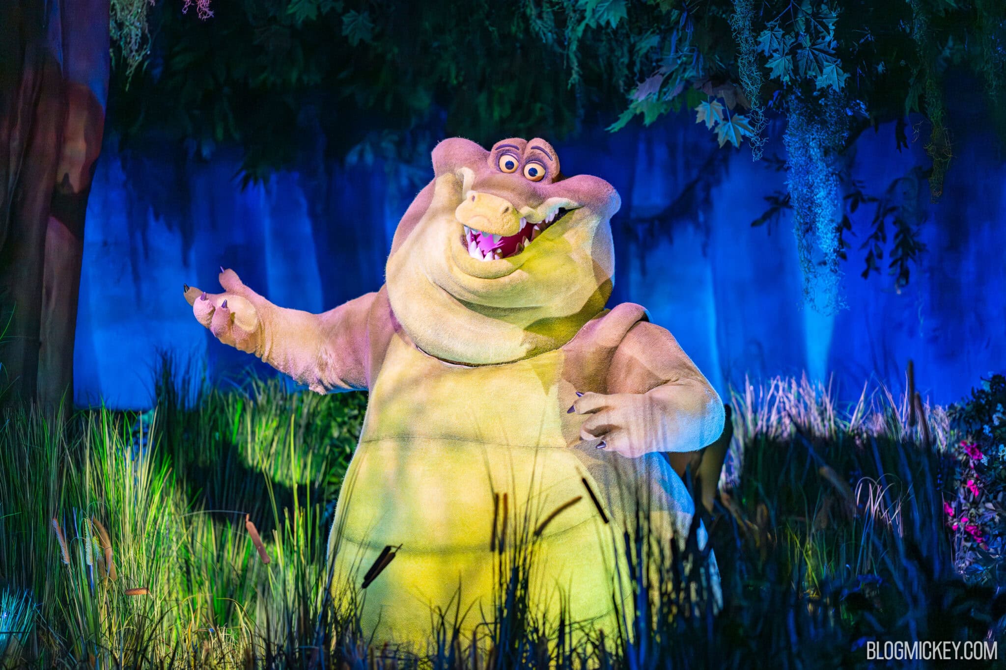 The Louis the alligator Audio-Animatronic standing among reeds with one arm outstretched and the other drawn in, lit blue against a swamp backdrop hung with Spanish moss