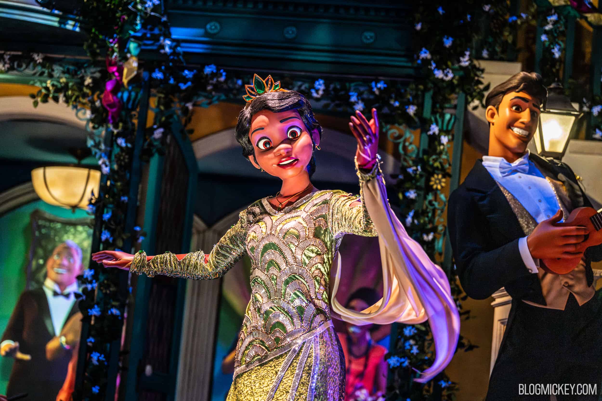 The Tiana Audio-Animatronic in the finale scene wearing a gold and green beaded gown and tiara with one arm raised, a tuxedoed figure holding a small stringed instrument at right