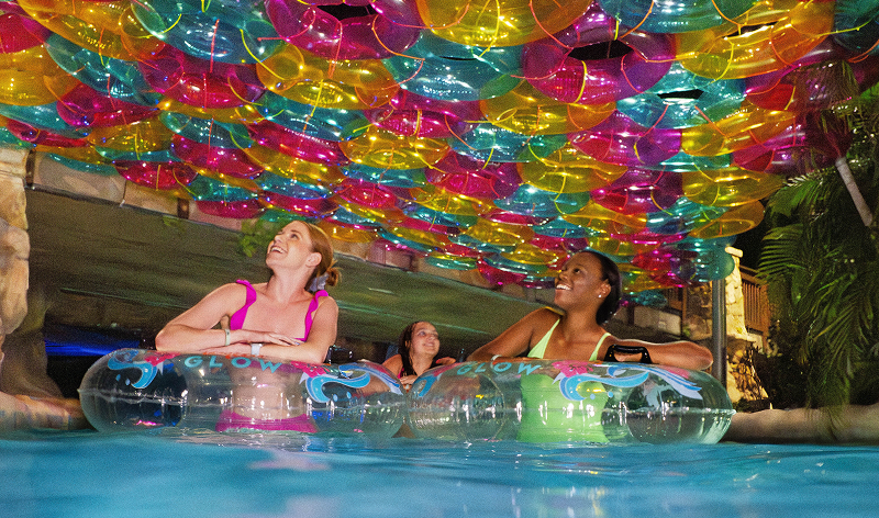 Three guests in a lit pool at night, two of them leaning on clear inner tubes printed with the word GLOW, beneath a dense canopy of multicoloured translucent orbs