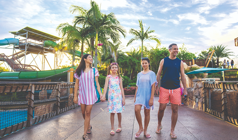 A family of four walking hand in hand along a concrete path at Aquatica, wood and wire mesh railings on either side, a green water slide and palm trees behind them in low sun