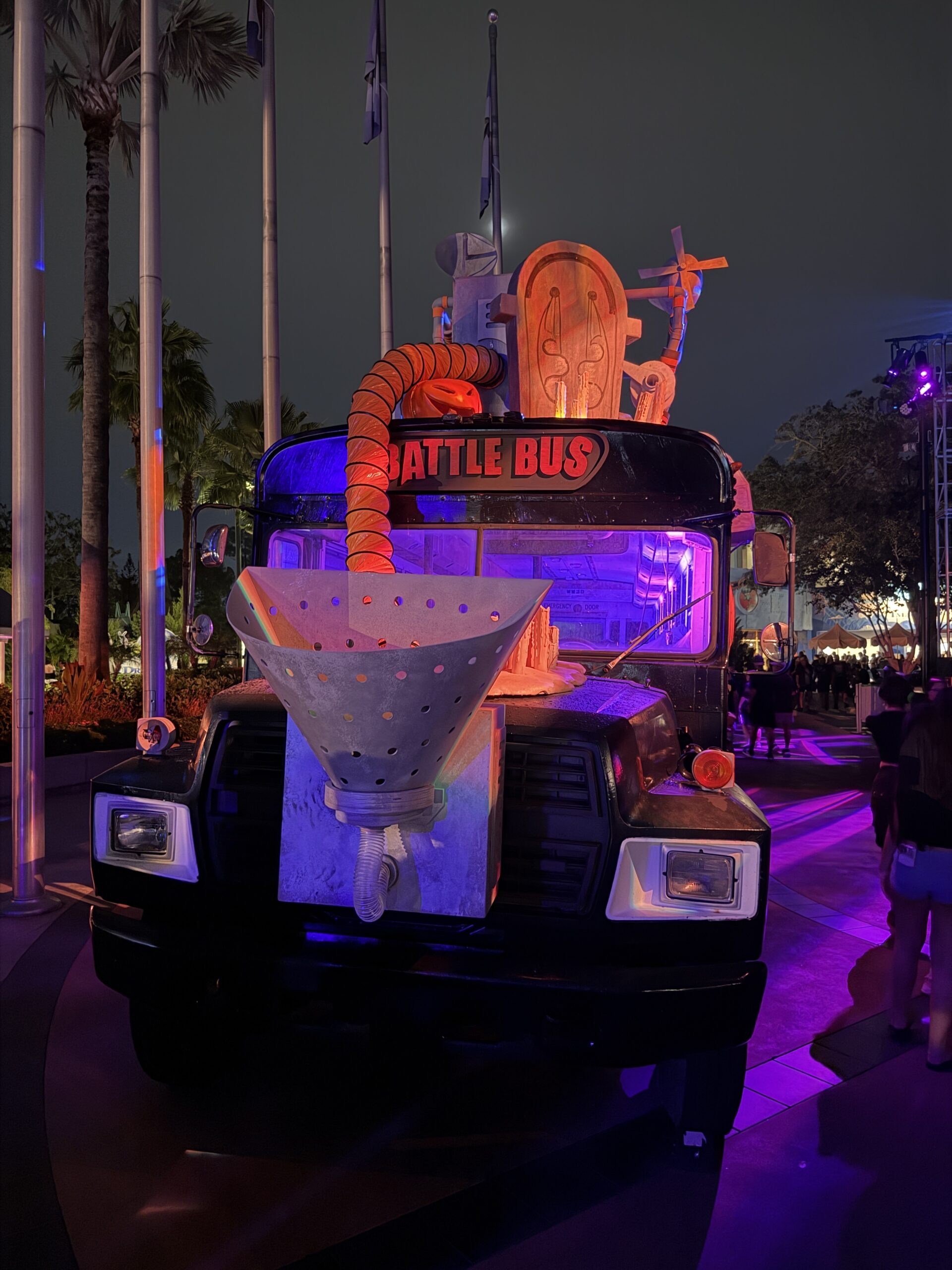 The front of a black school bus lettered BATTLE BUS in red above the windshield, with a large pale funnel on the grille, orange ducting, and purple lit windows, palm trees behind