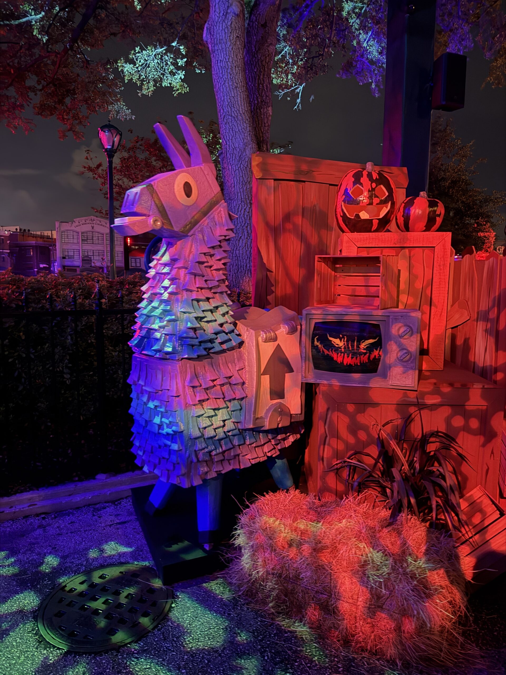 A large loot llama pinata lit in blue and pink standing beside stacked wooden crates, a carved jack-o-lantern, a retro television showing a grinning mouth, and straw bales