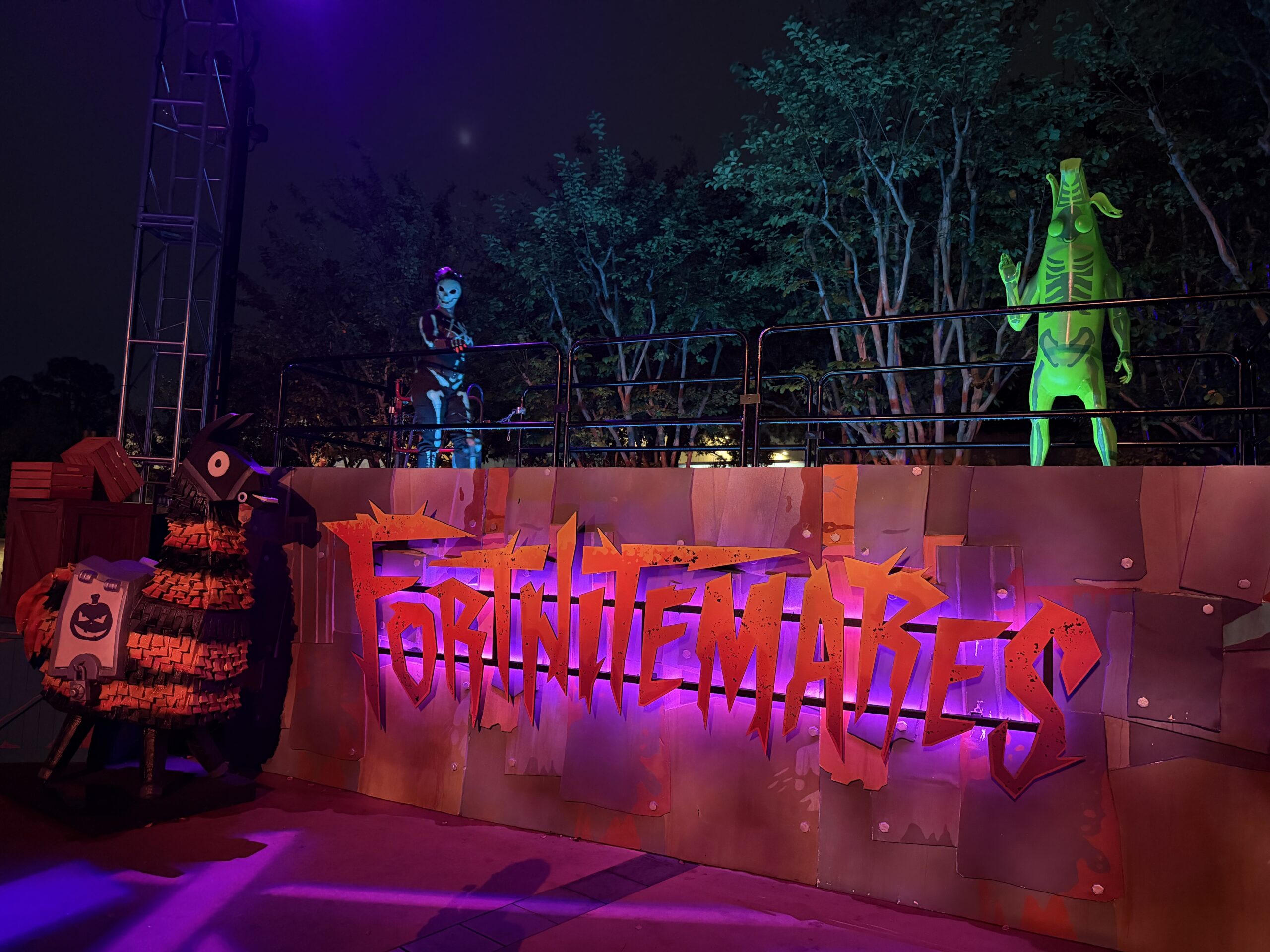 A scareactor in a skull mask and a tall green skeleton-printed banana figure standing on a raised platform behind railings, above an orange FORTNITEMARES sign on a patched metal wall, with a dark loot llama pinata at left
