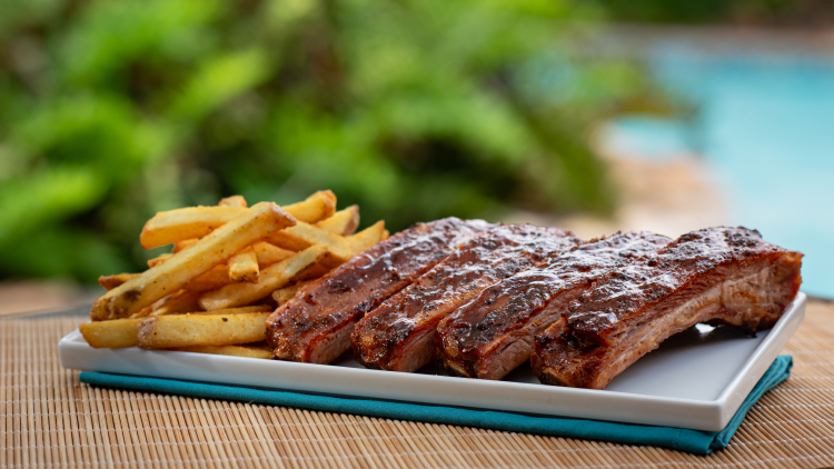 A rack of barbecue ribs and a pile of french fries on a white rectangular plate set on a bamboo mat, with greenery and a blue pool behind