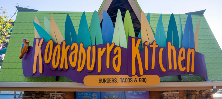 The Kookaburra Kitchen sign, orange script lettering on a purple banner with multicoloured leaf shapes above and a kookaburra bird figure at left, over an orange oval reading BURGERS, TACOS & BBQ