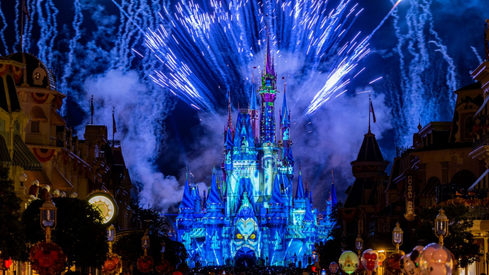 Cinderella Castle lit in blue projections with white fireworks fanning out overhead, framed by Main Street buildings