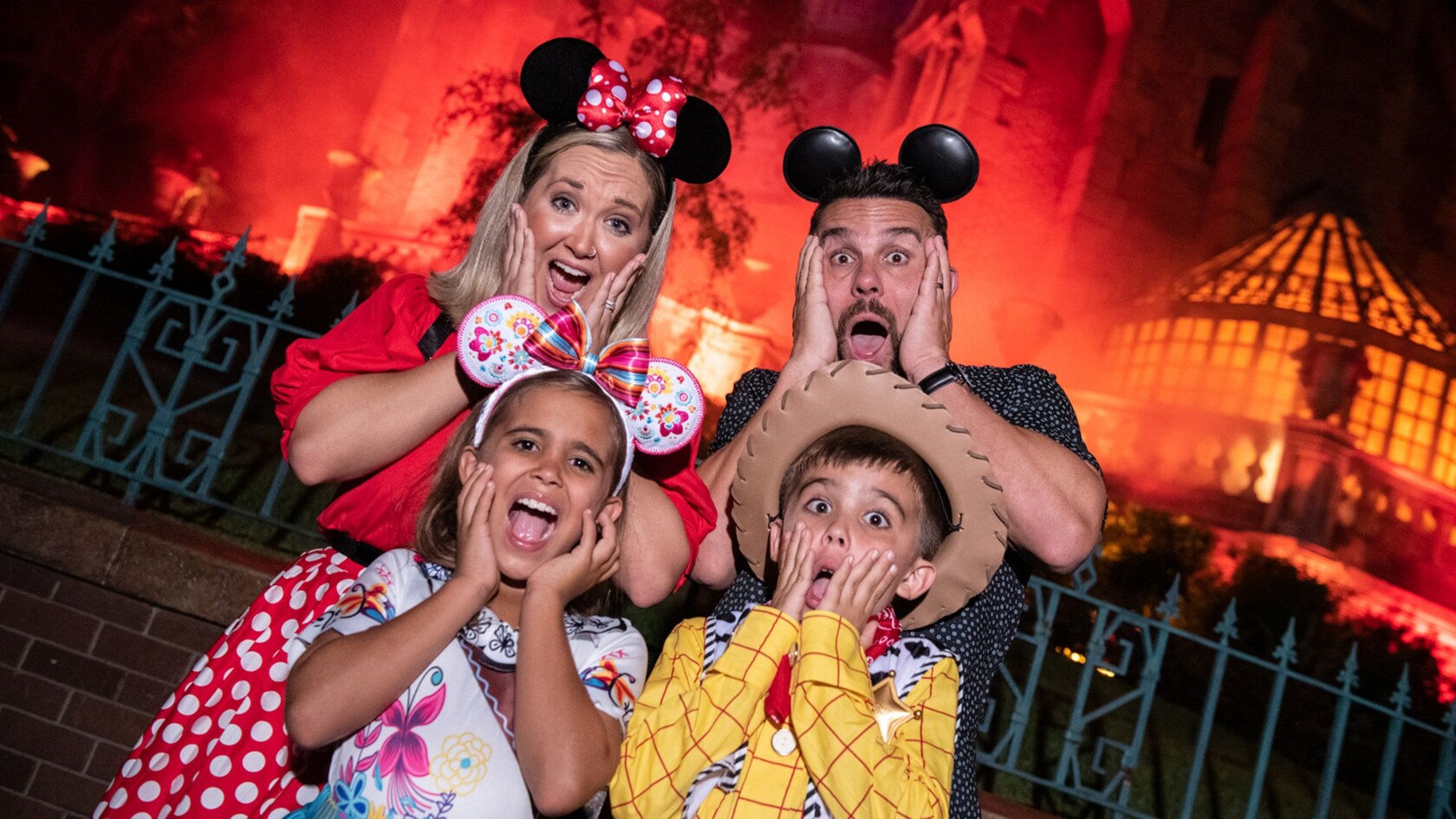 Two adults and a girl in Mickey and Minnie ears plus a boy in a Woody costume, all making mock-frightened faces in front of a red-lit Haunted Mansion at night