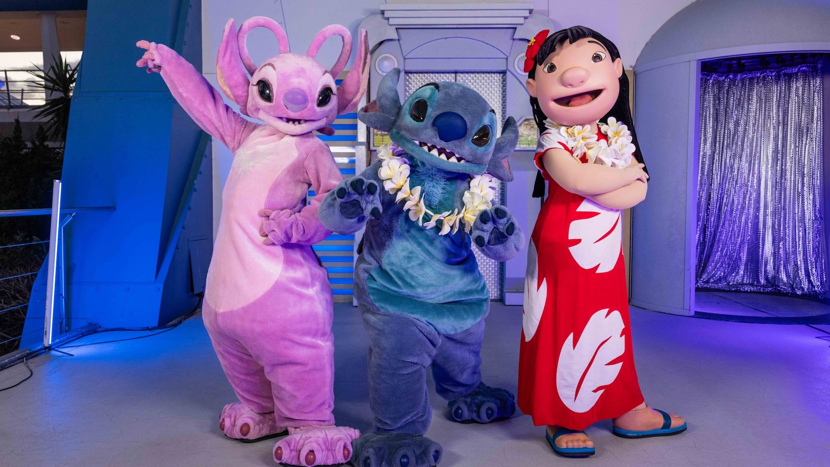 Angel in pink, Stitch in blue wearing a flower lei, and Lilo in a red dress posing together in a blue-lit setting
