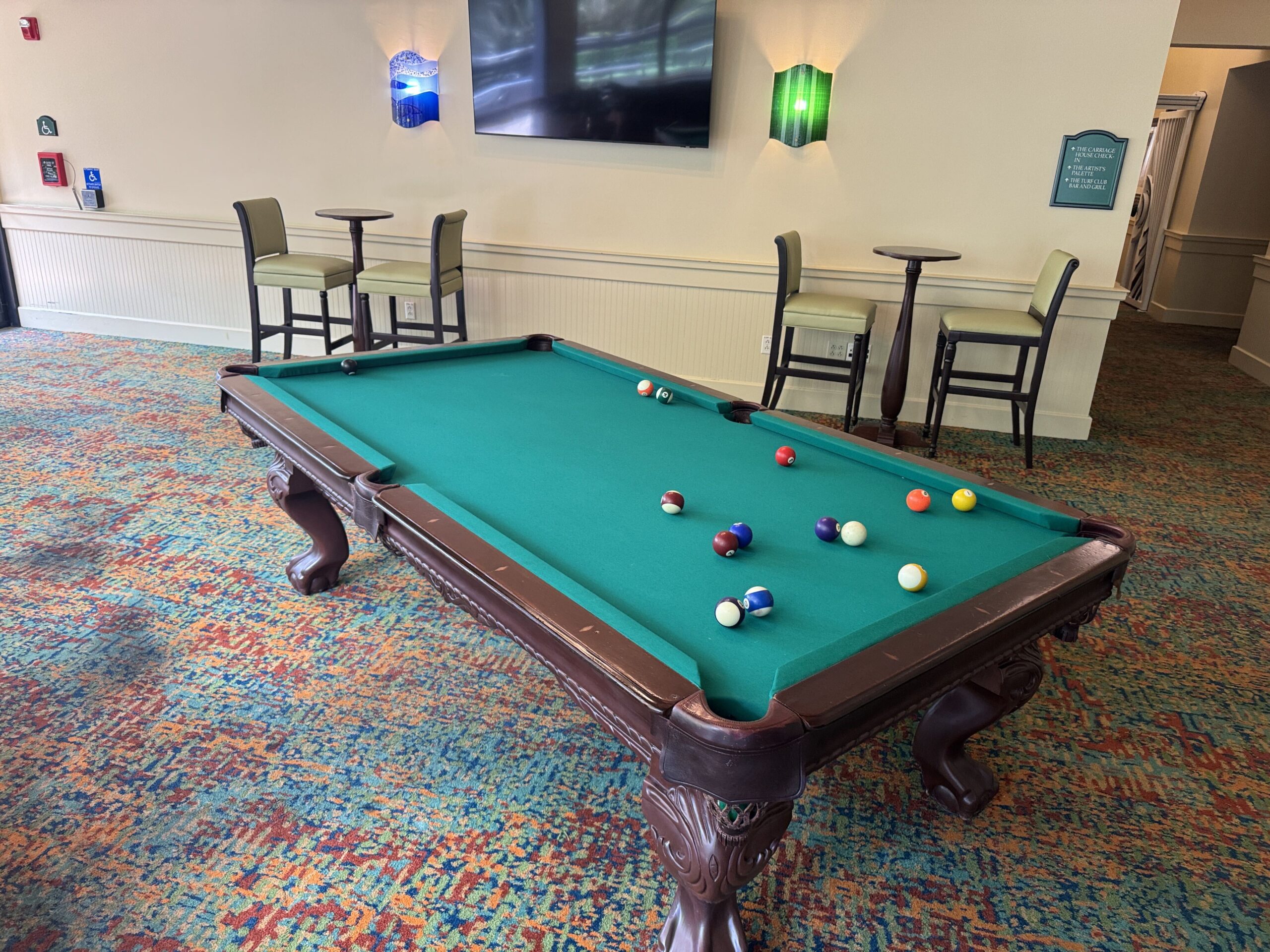 A green felt pool table with balls scattered mid-game on a multicolored carpet, with olive barstools, a wall-mounted television and a green directional sign behind it